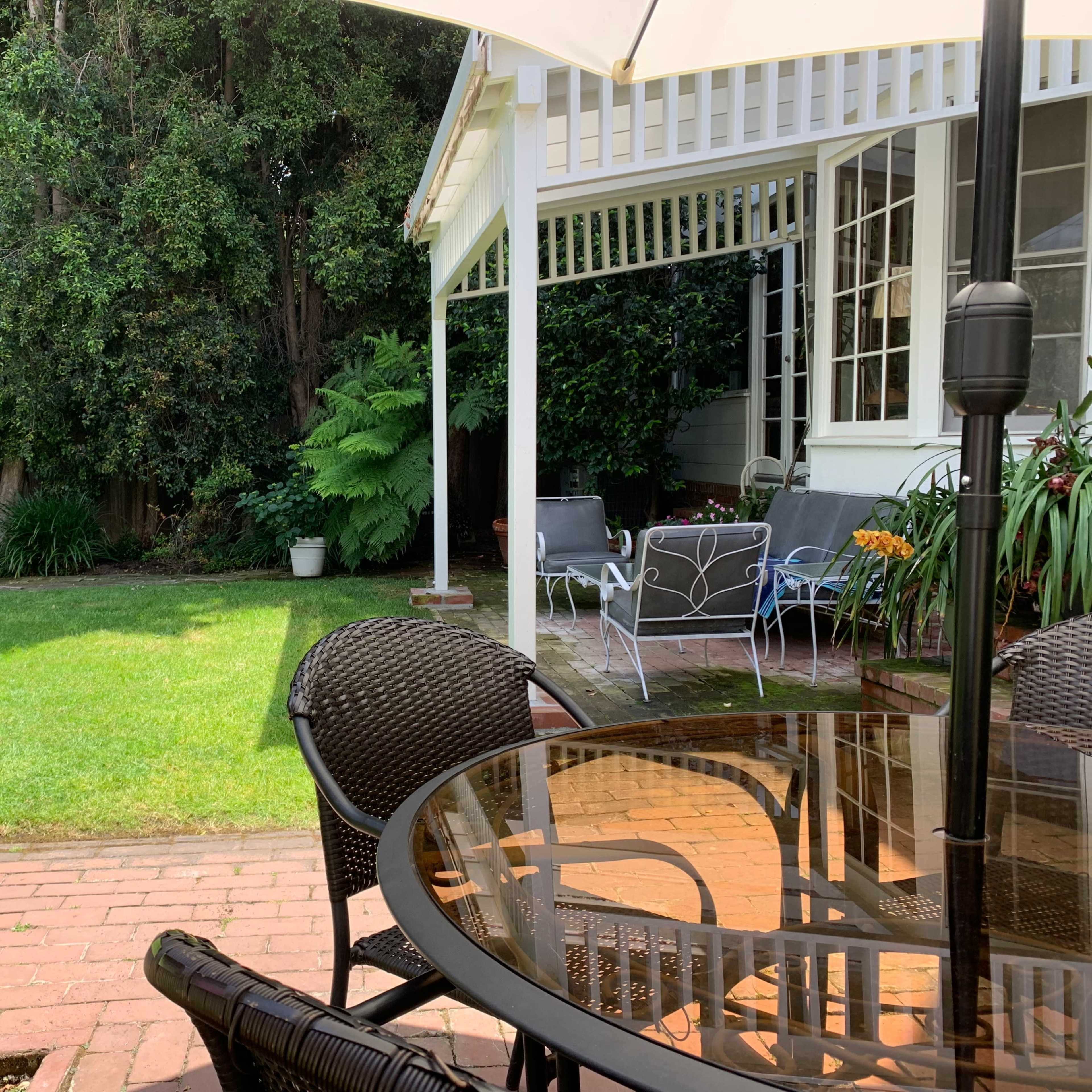 A patio table with chairs sits in front of a well-maintained garden featuring green grass and ornamental plants, under a shaded umbrella.