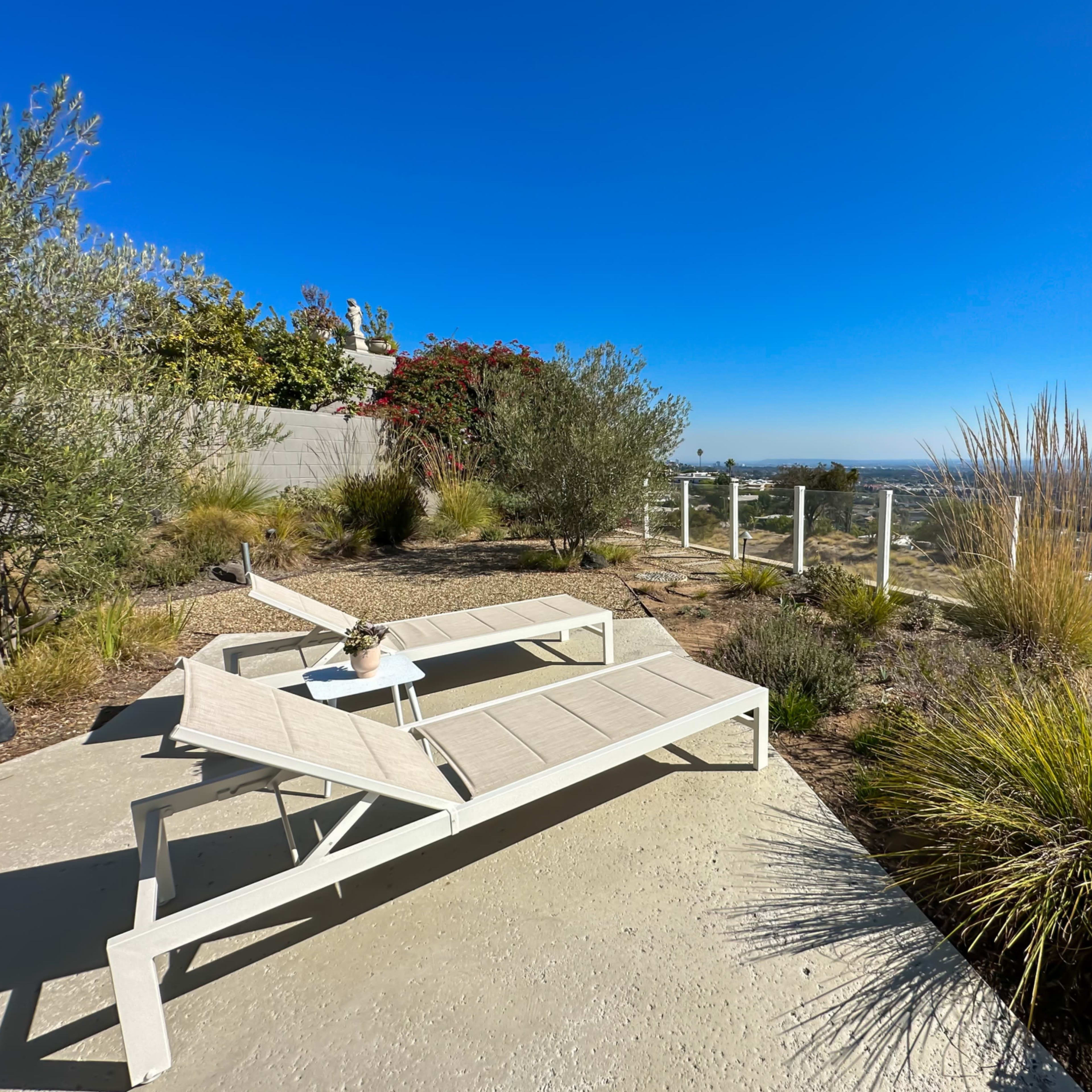 A pair of white lounge chairs sit on a concrete patio overlooking a wide view of a landscape with shrubs and grass under a clear blue sky.