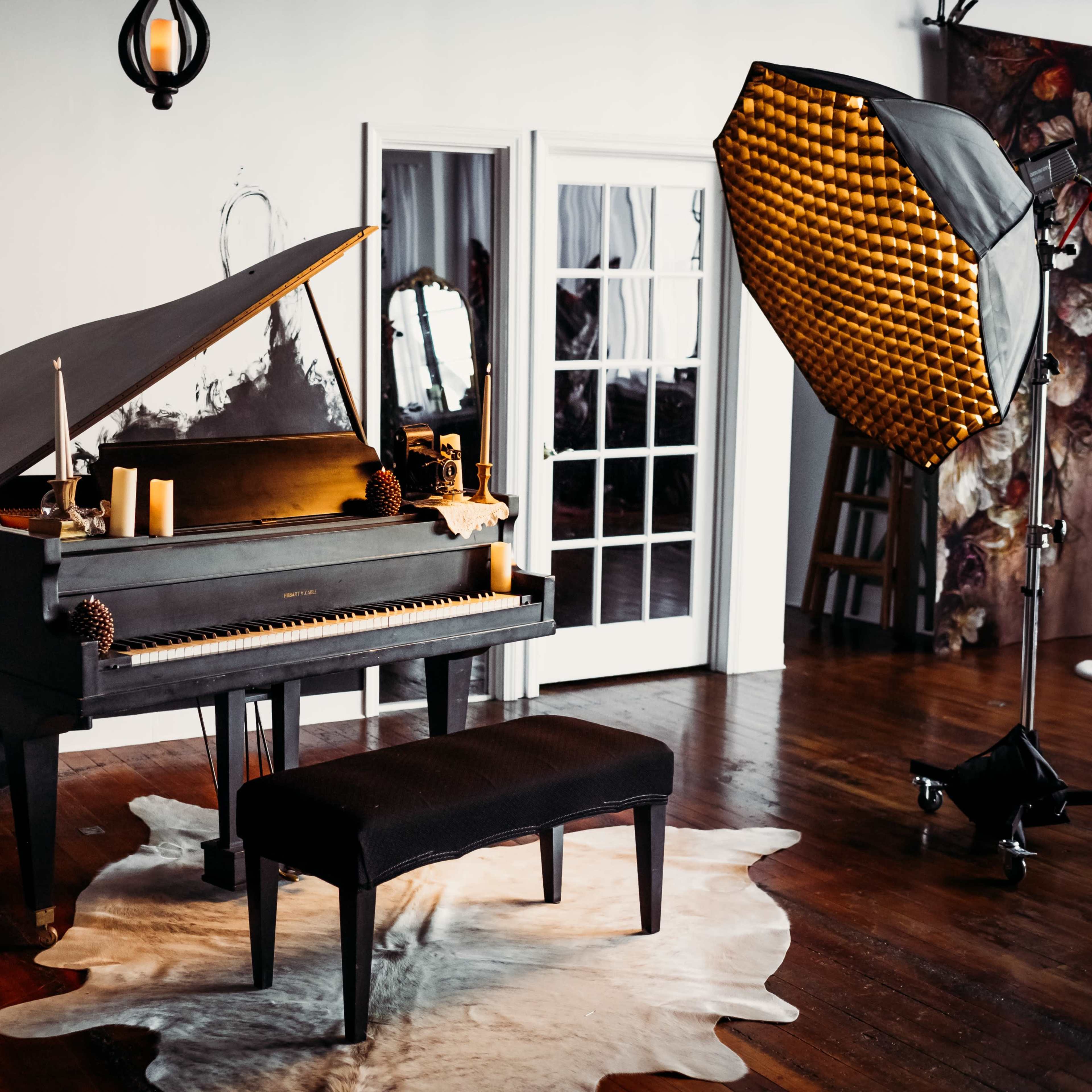 A black upright piano with a matching bench is positioned on a cowhide rug in a well-lit room featuring a large window and a photography light setup.