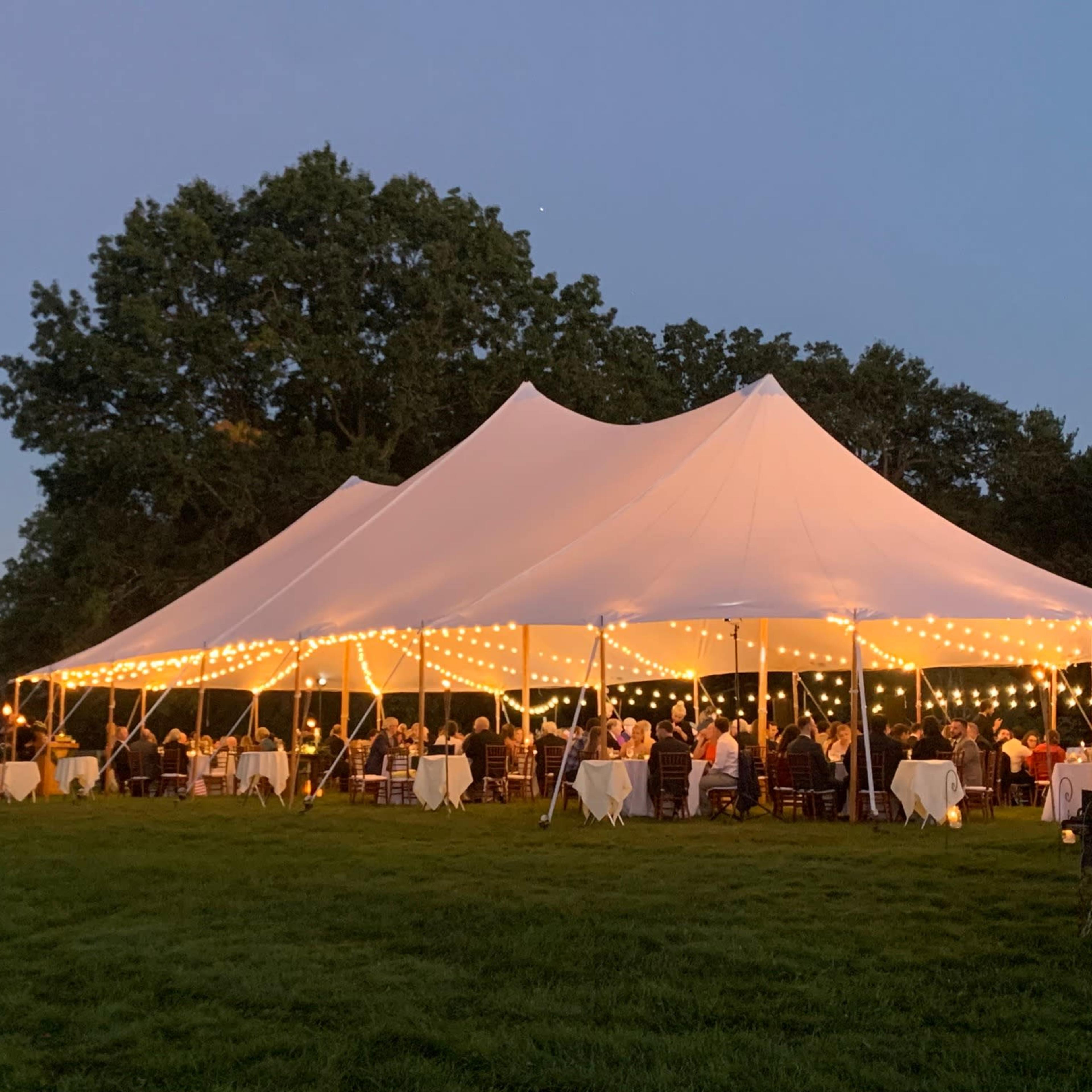 A large white tent decorated with string lights hosts an outdoor gathering on a grassy area near a tree line, with a bright moon visible in the sky.