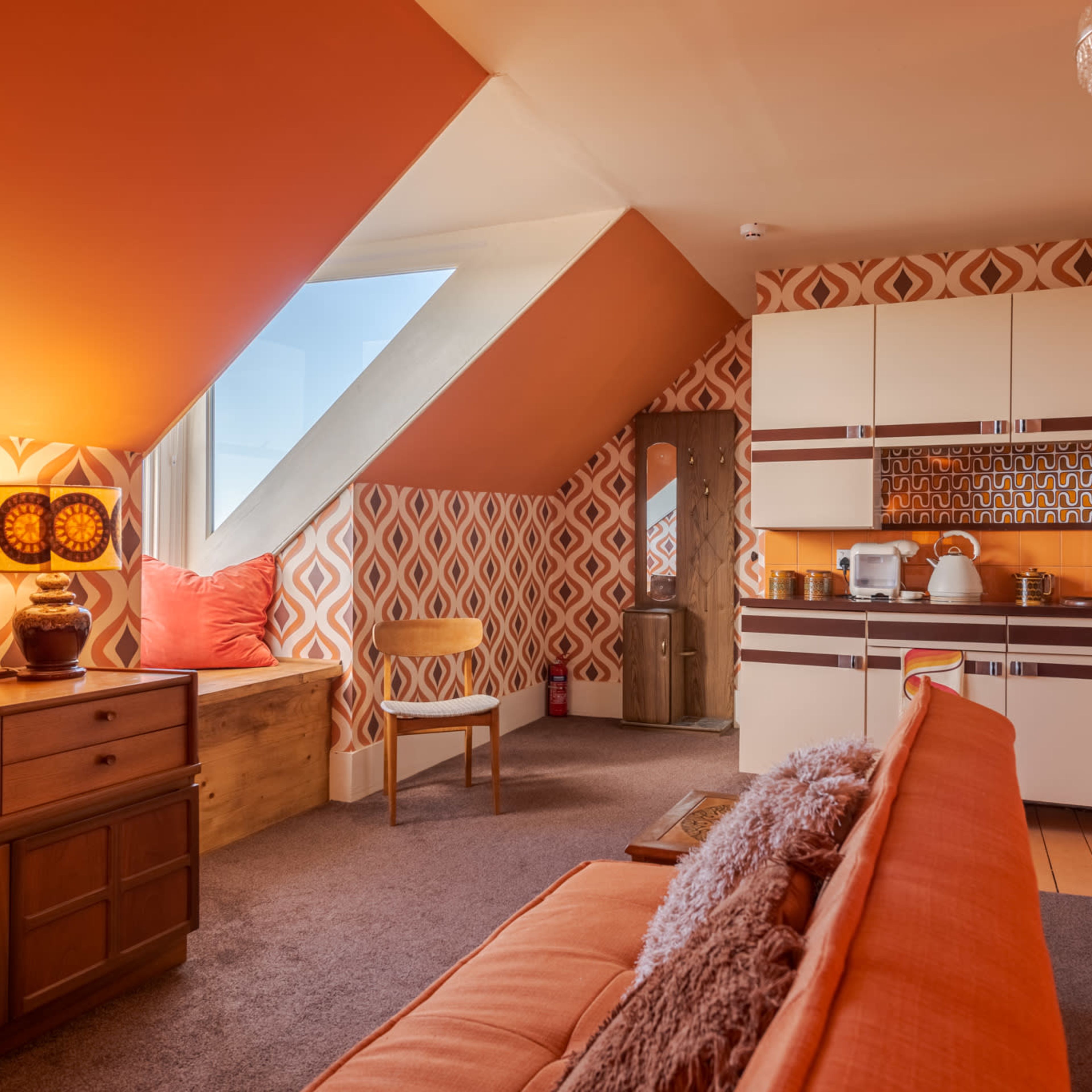 A penthouse room features orange geometric wallpaper, a kitchen area with retro cabinets, and a cozy seating arrangement.