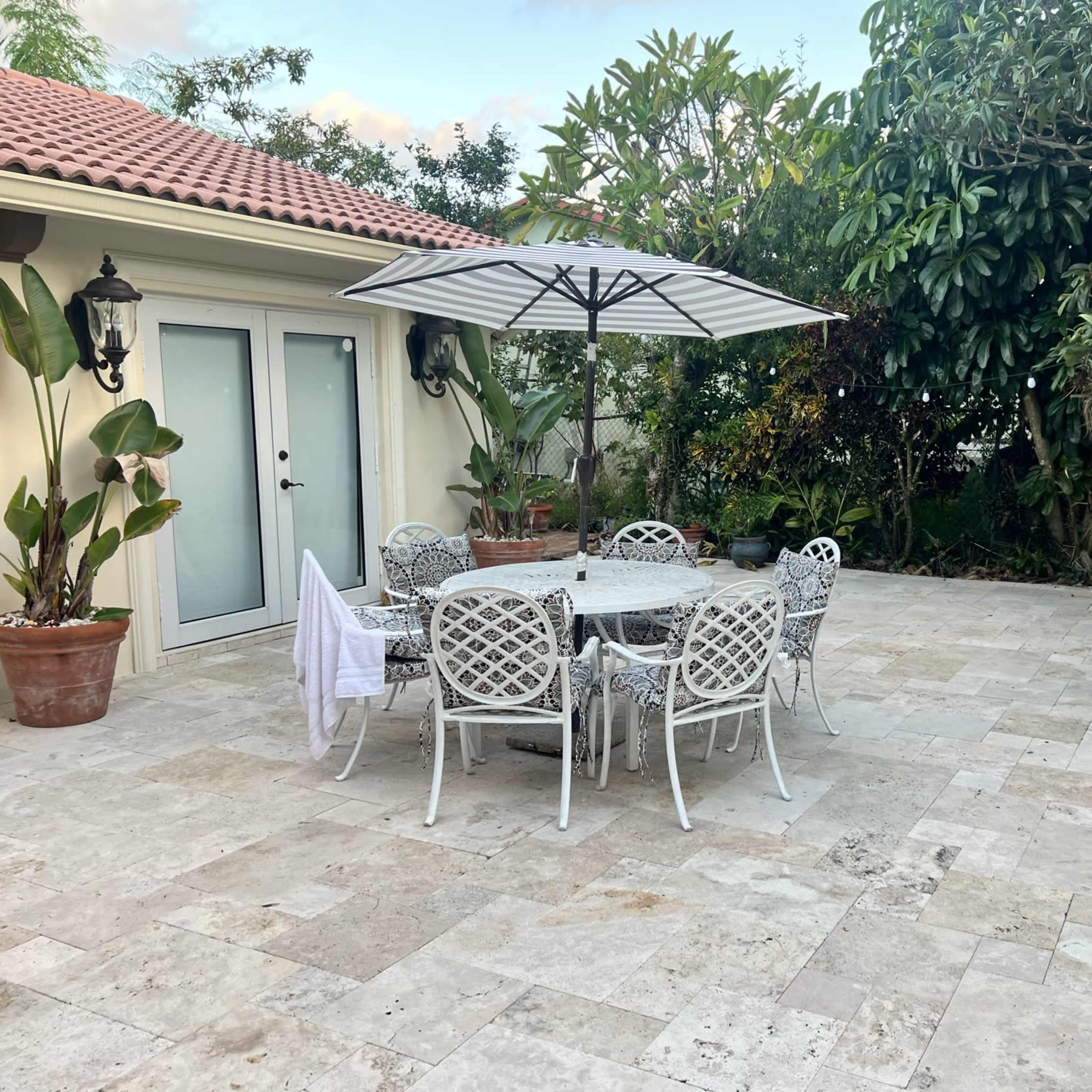 A stone patio features a round dining table surrounded by white metal chairs, shaded by a large umbrella, with lush greenery in the background.