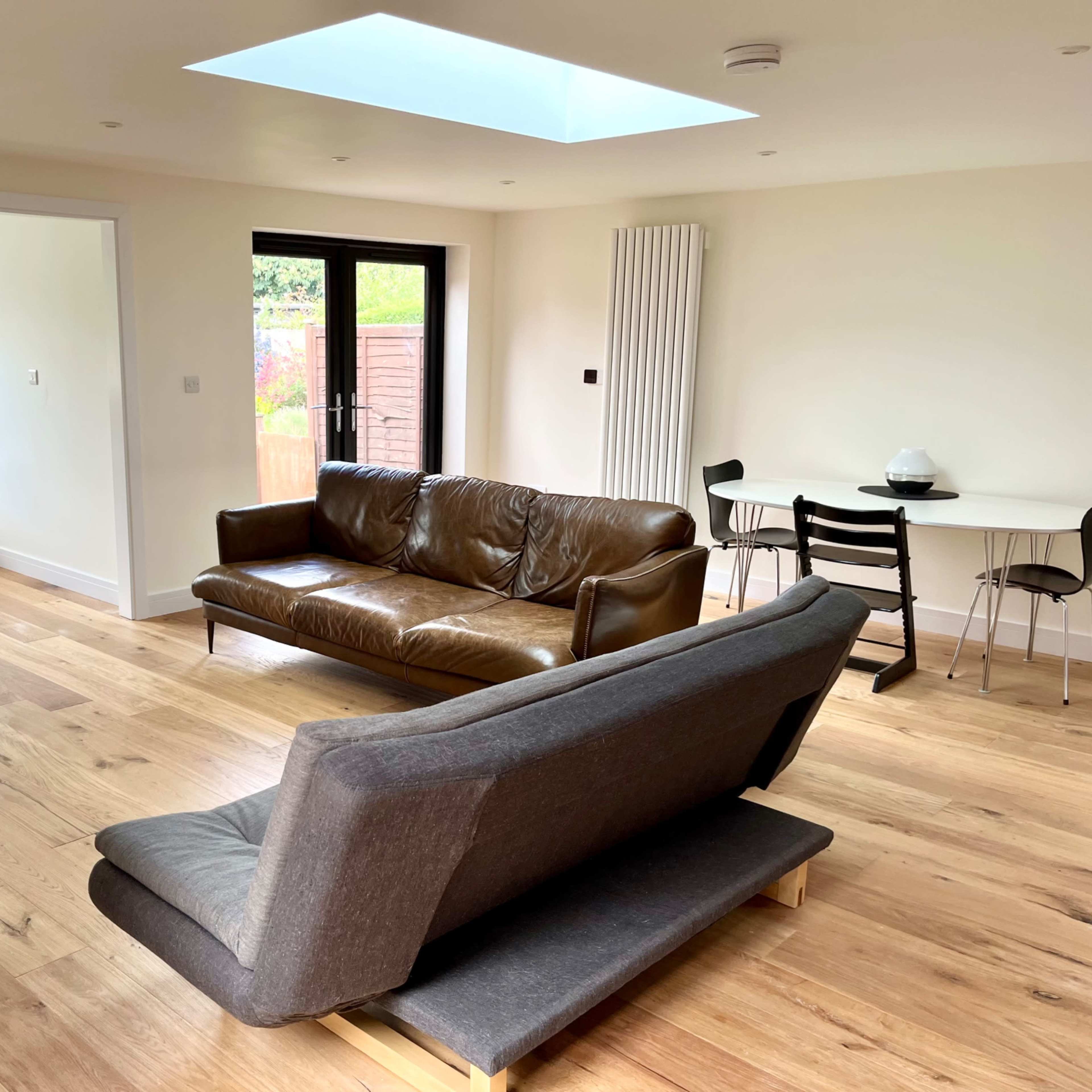 A modern, open-concept living space with a brown leather sofa, a gray couch, a circular dining table, and large windows allowing natural light to enter.
