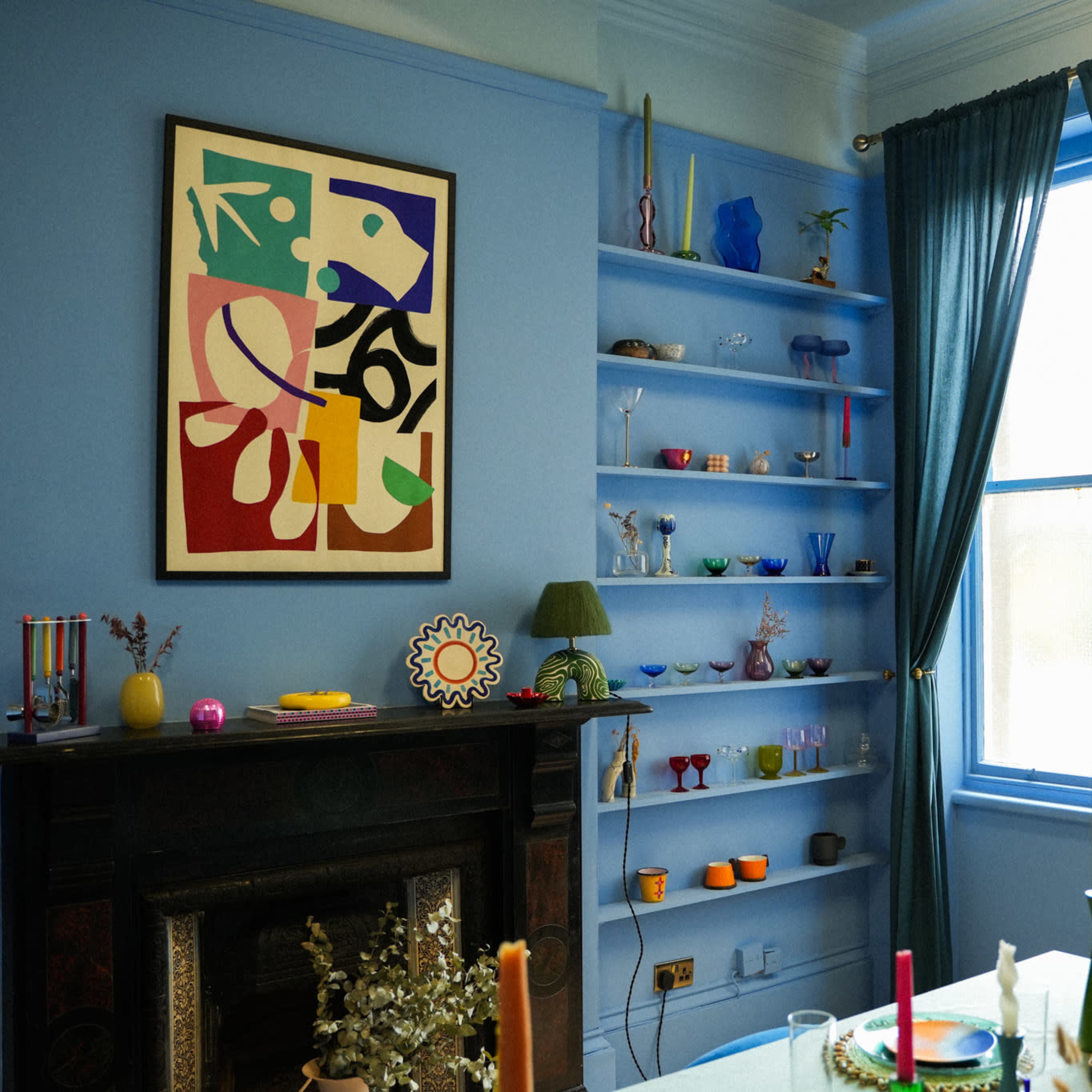 The room features a blue-painted wall adorned with colorful artwork and shelves displaying various decorative items, alongside a table set with candles and a vase of flowers.