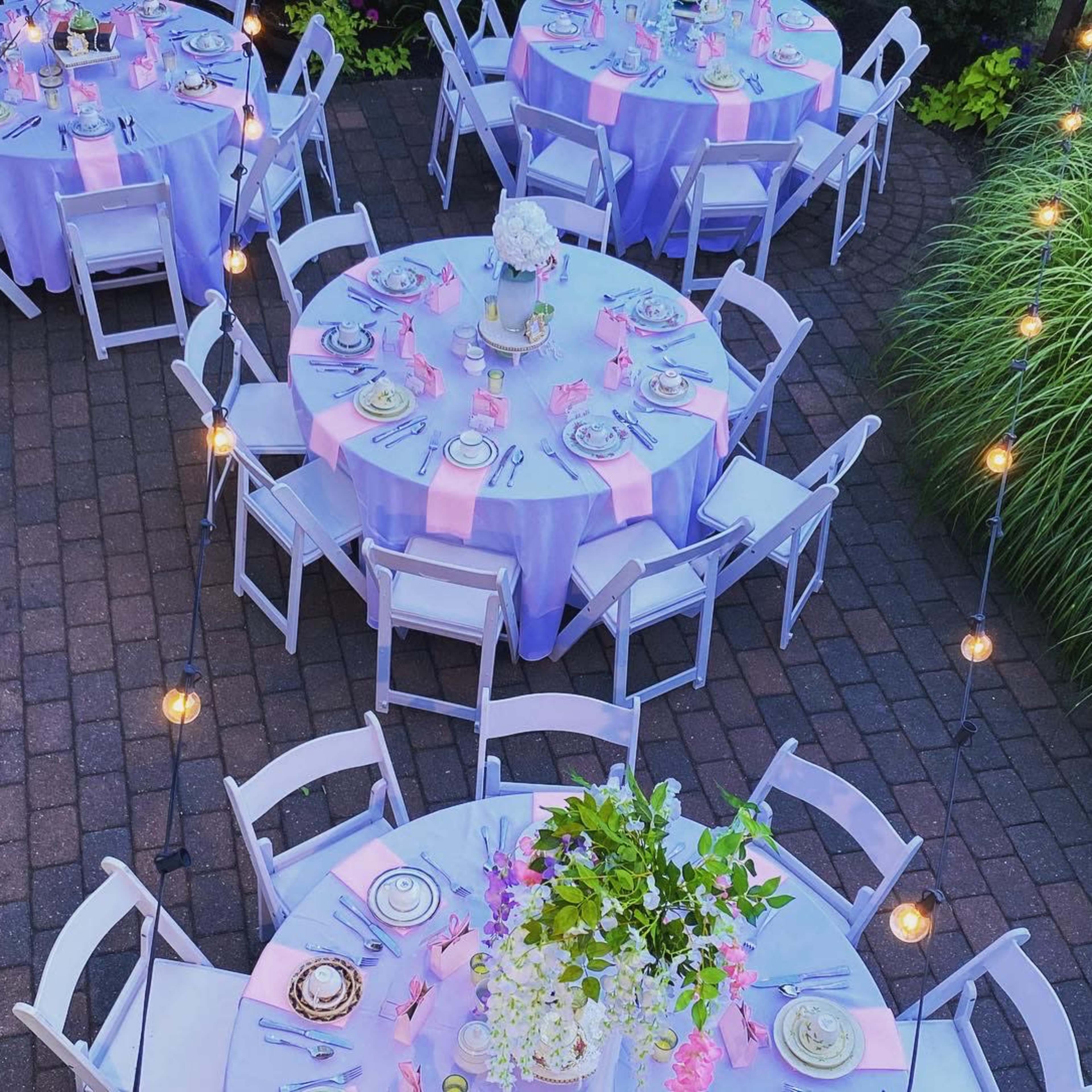 An outdoor dining setup with round tables covered in lavender tablecloths, surrounded by white chairs and adorned with floral centerpieces, all under hanging string lights.
