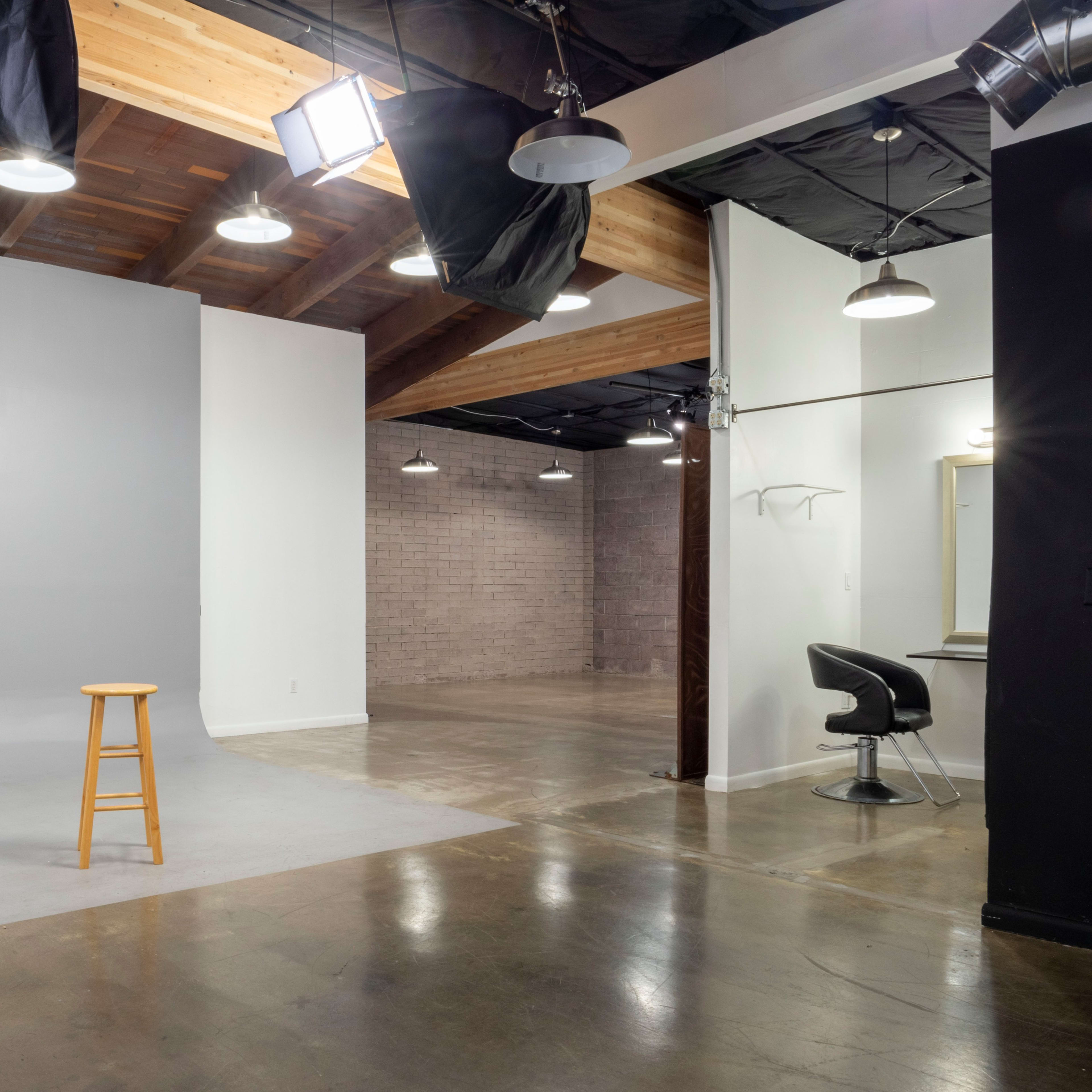 A spacious studio with a gray backdrop, a wooden stool in front, and a separate area featuring a black chair and a mirror.