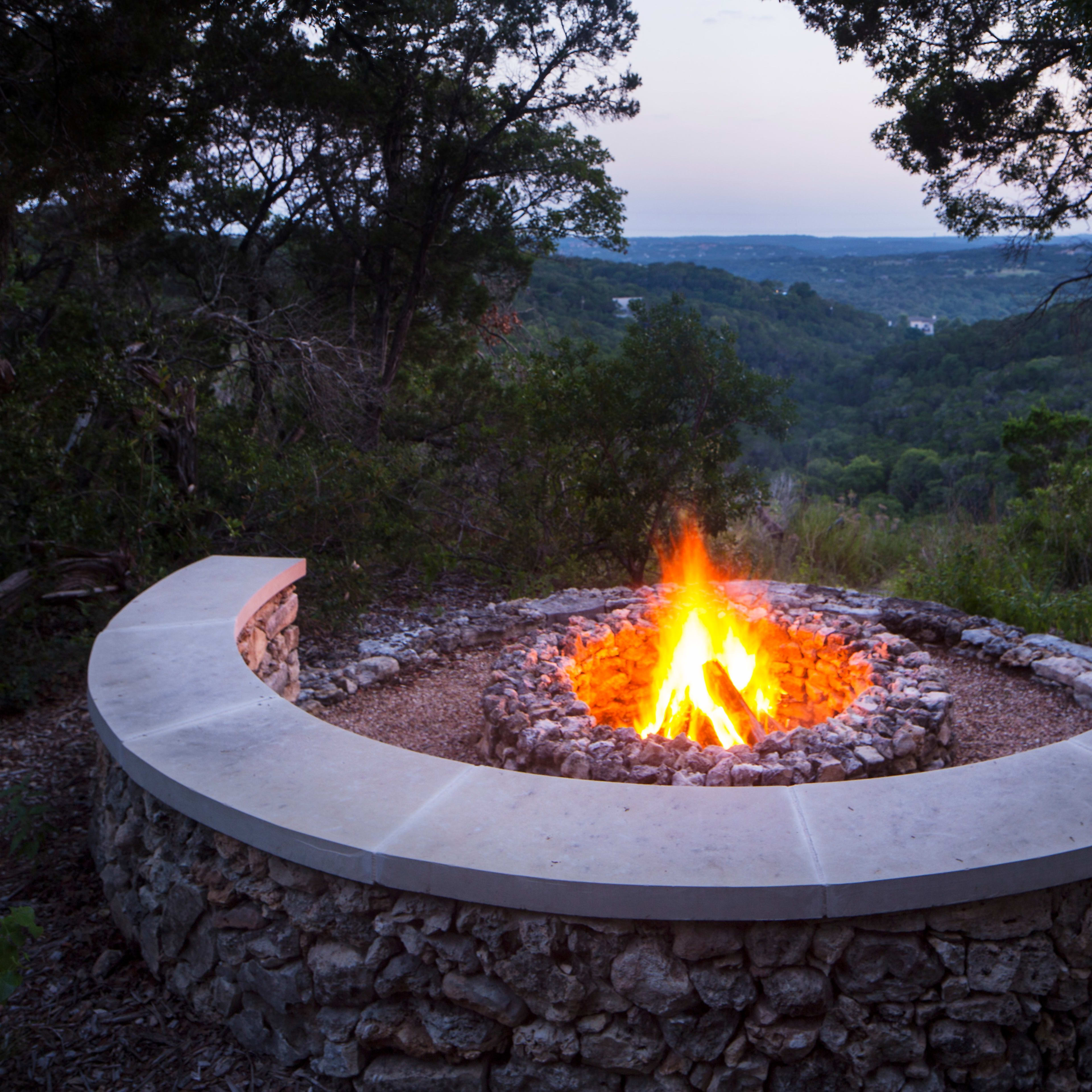 A circular stone fire pit with a vibrant flame is set against a backdrop of a lush, green hillside and a twilight sky.