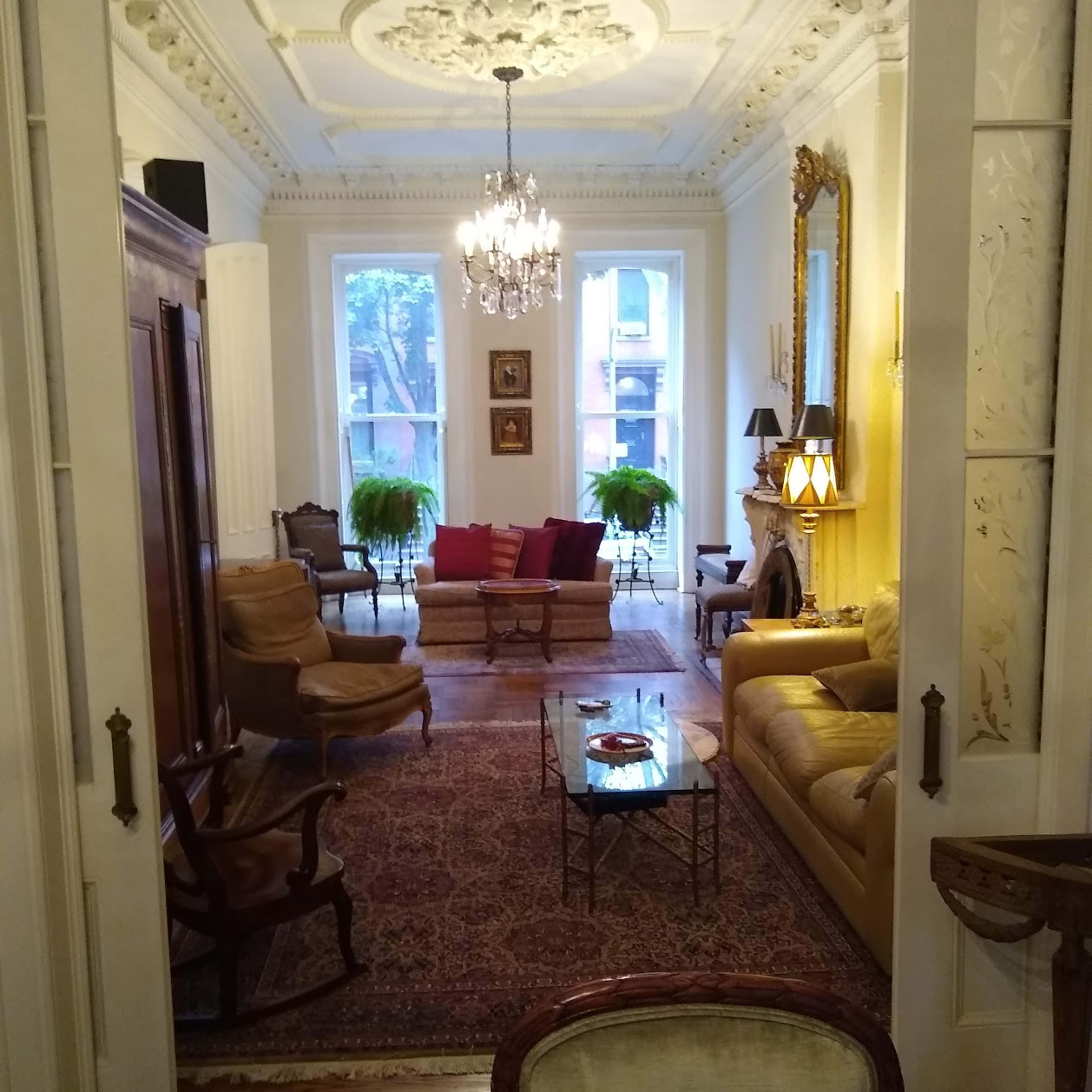 A stylish living room features a chandelier, ornate ceiling, and arranged seating around a coffee table, with large windows allowing natural light.