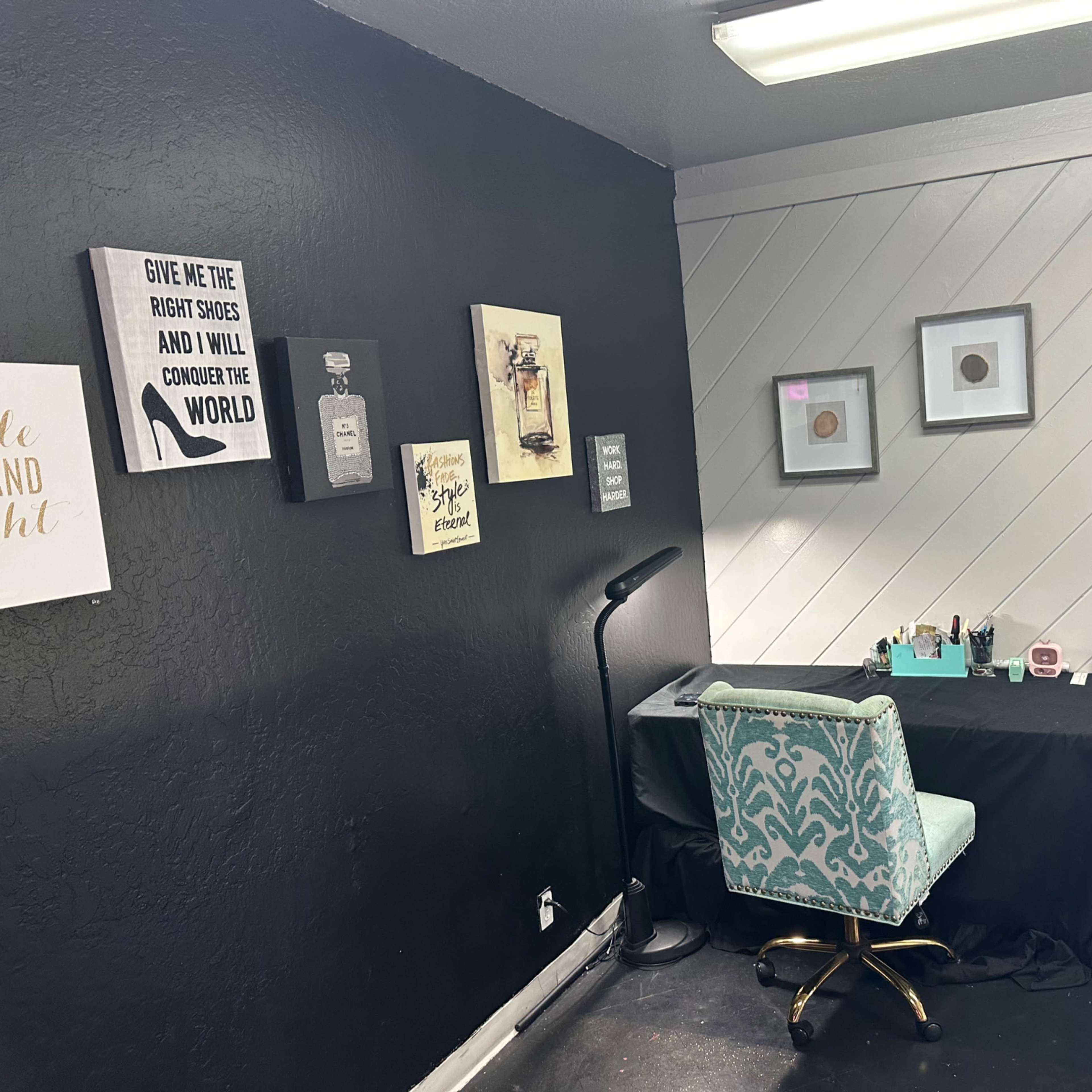 A small, neatly arranged workspace with a black wall and framed artwork, featuring a desk with a chair, lamp, and various supplies.
