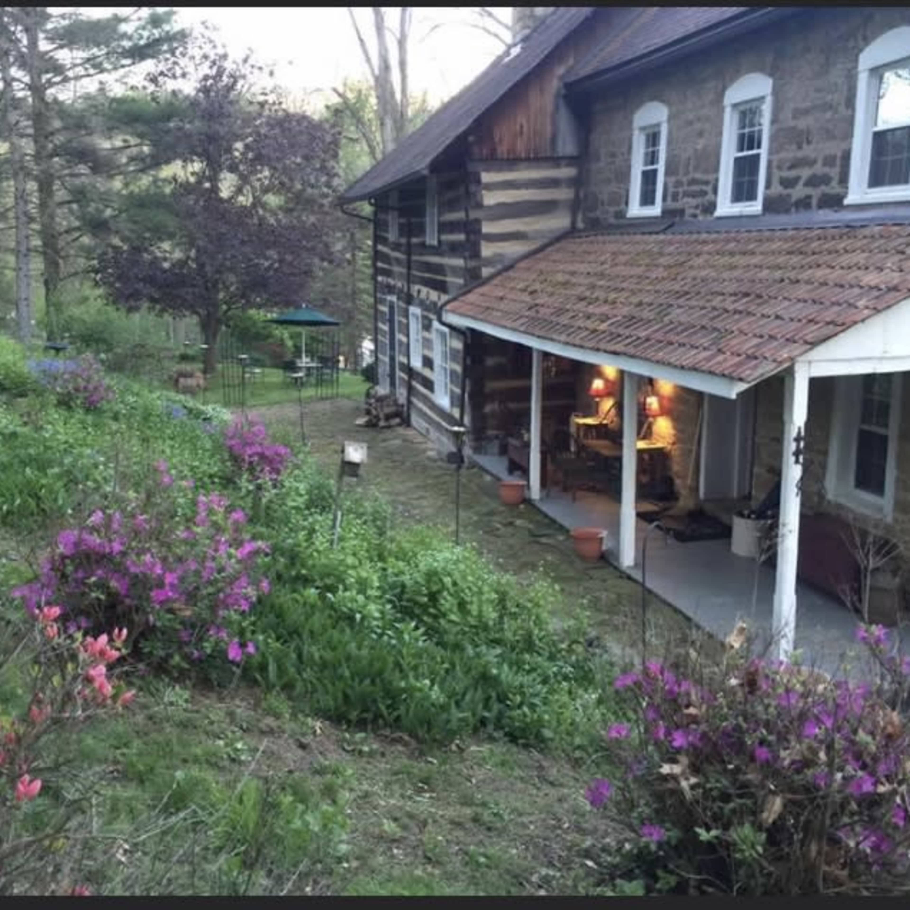 A stone house with a wooden porch is surrounded by blooming azaleas and a garden in a wooded area.