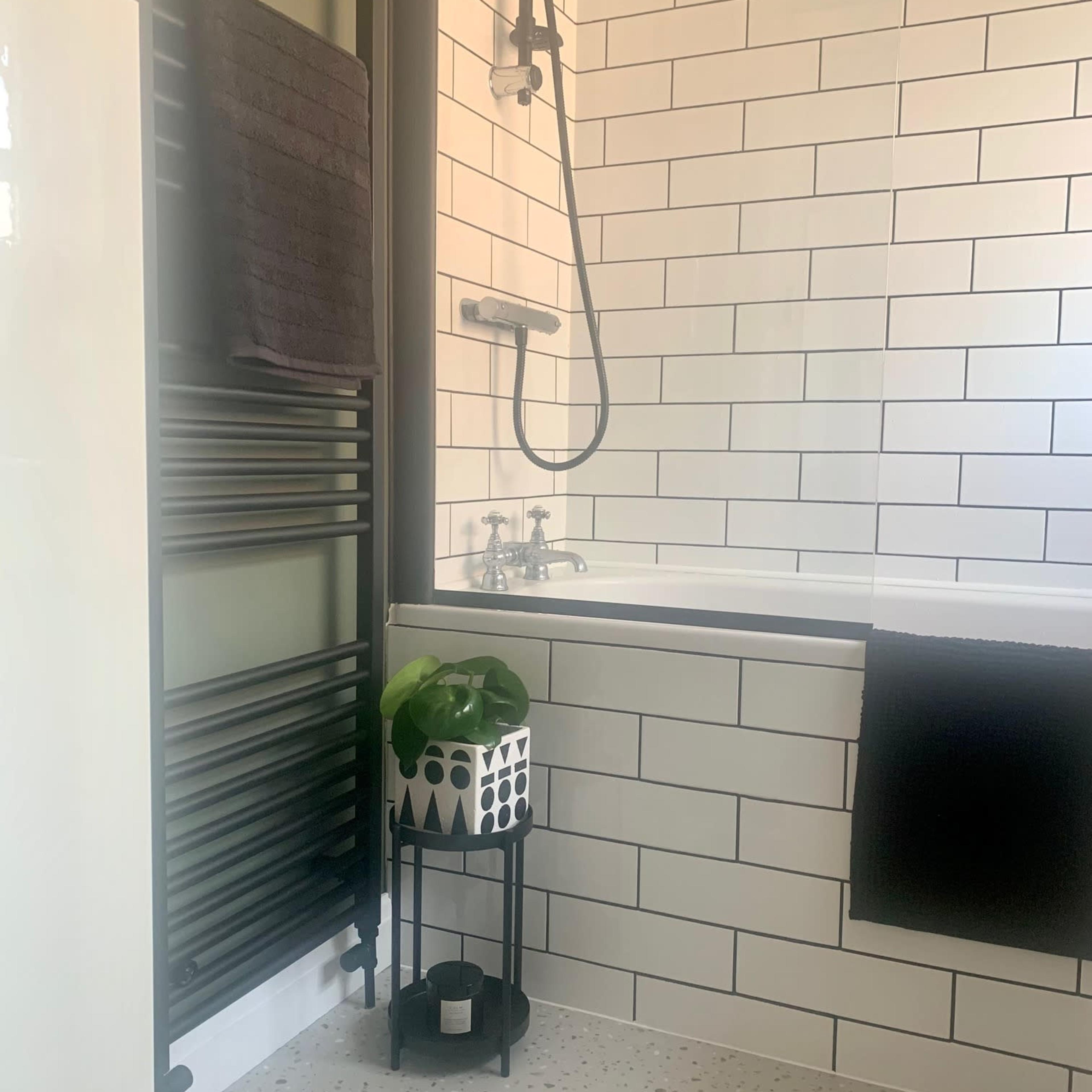 A modern bathroom with a bathtub, showerhead, a towel rack, and a potted plant on a stand beside the tub.