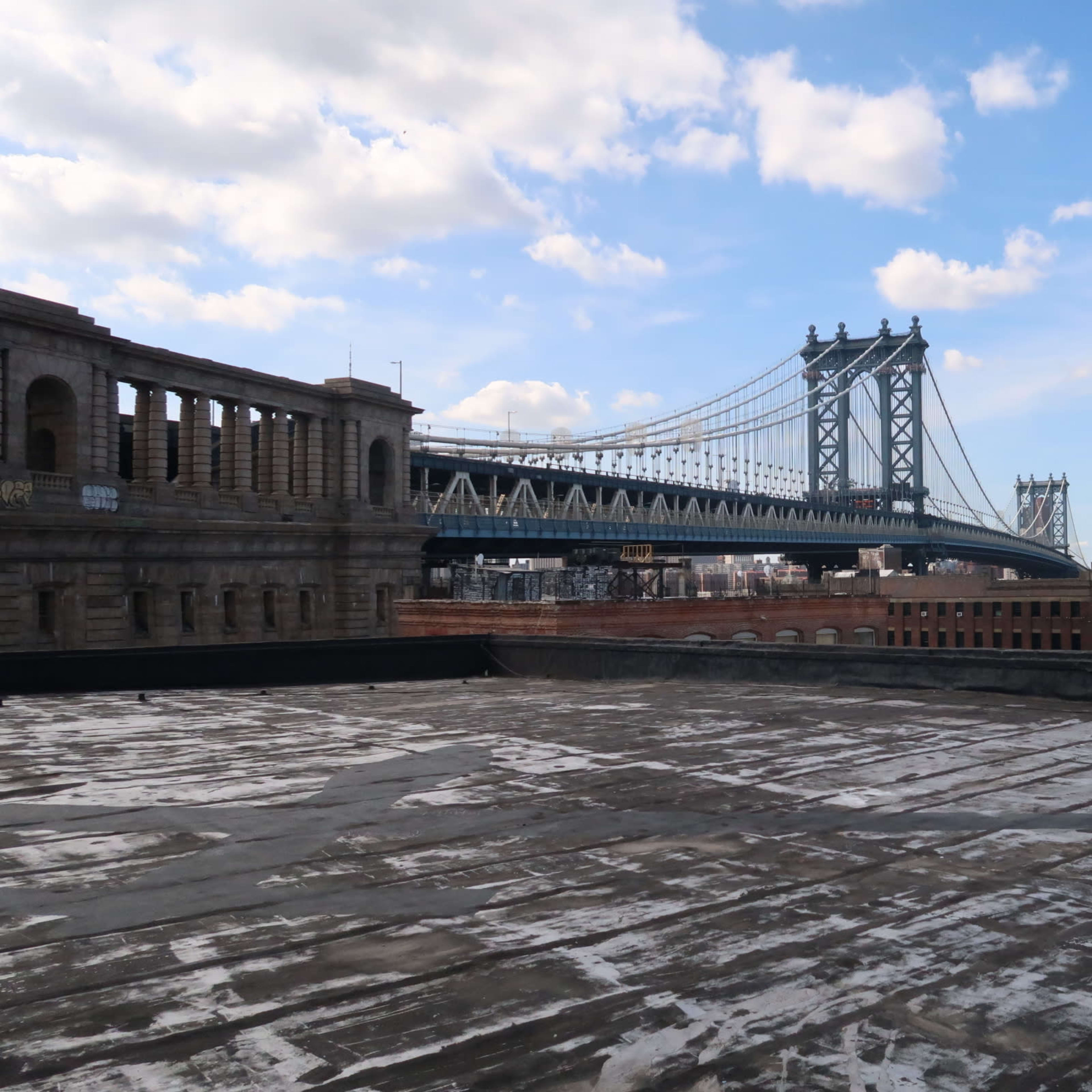 A rooftop view of the Manhattan Bridge with buildings in the background under a partly cloudy sky.