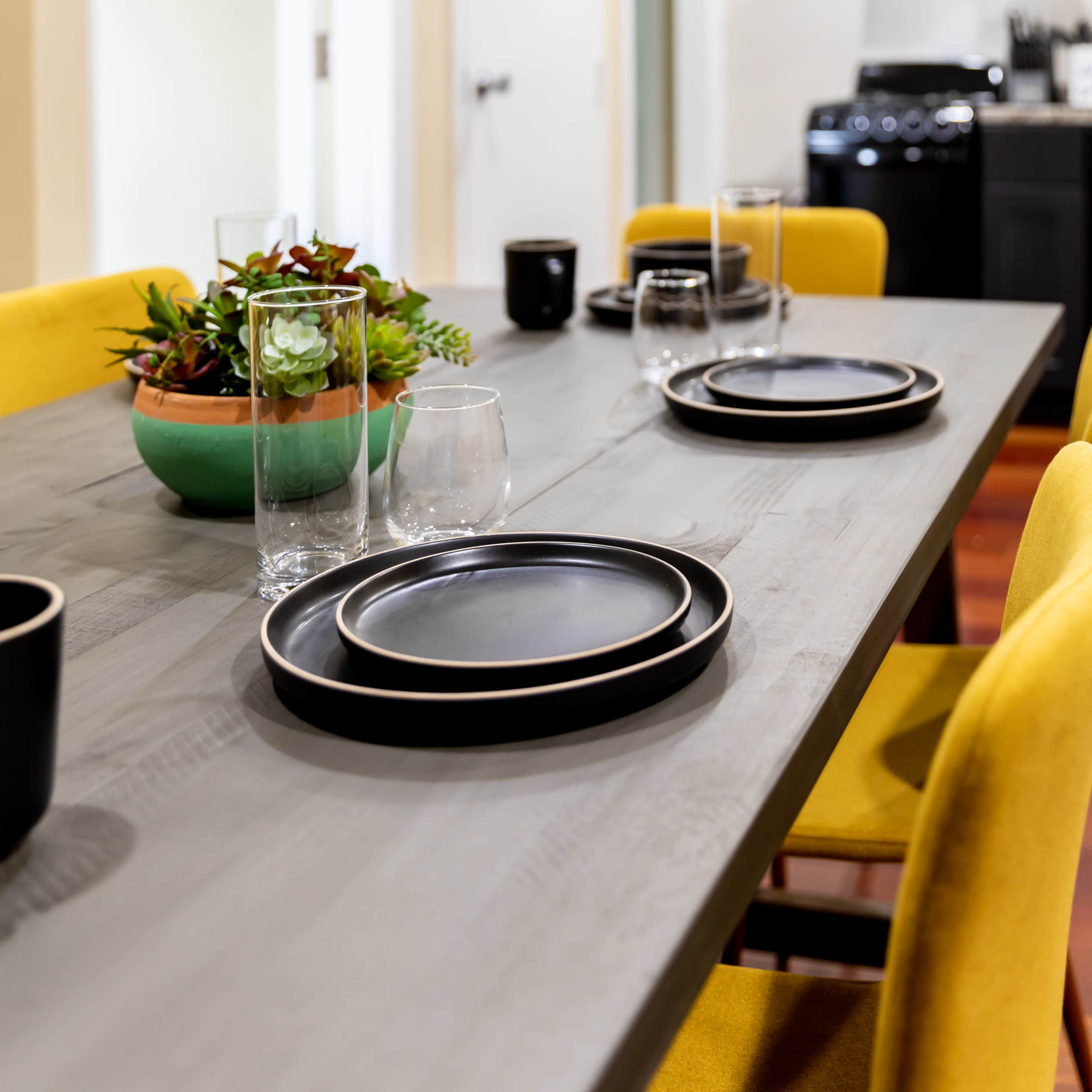 A dining table is set with black plates and glasses, surrounded by yellow chairs and a potted plant in a turquoise bowl, while a kitchen area is visible in the background.