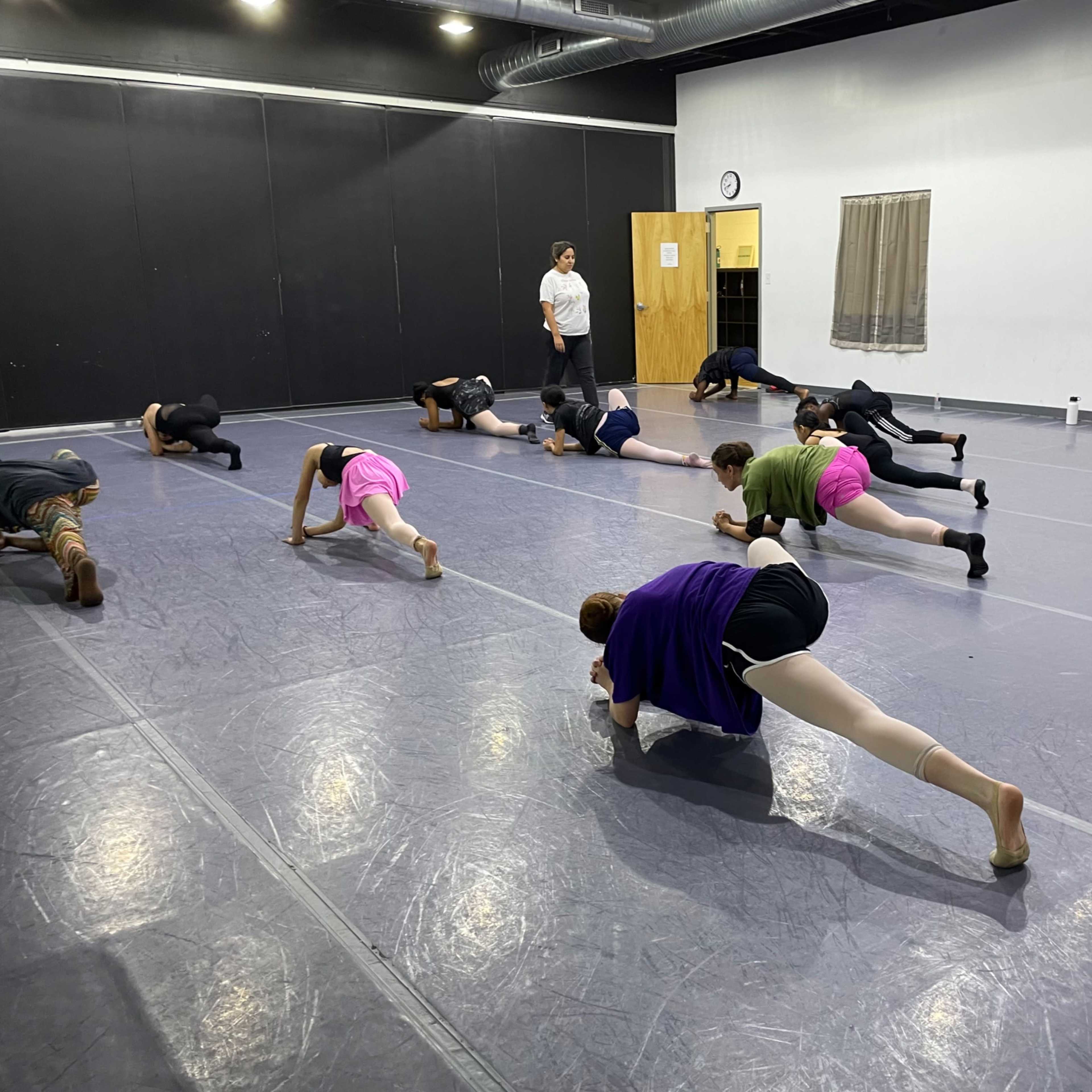 A group of dancers stretches on the floor of a rehearsal studio while an instructor observes in the background.