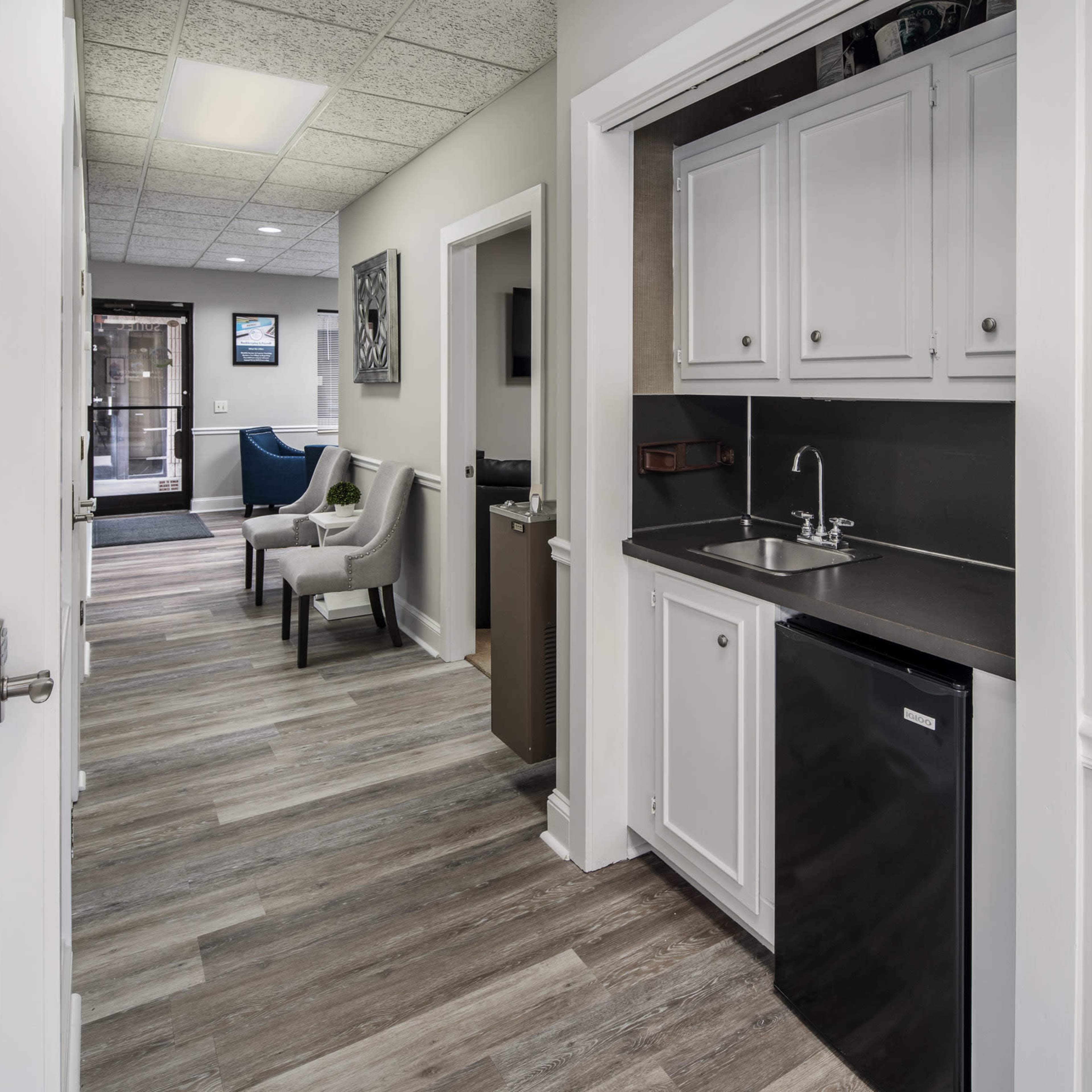 A hallway with a kitchenette on one side, featuring a sink and small refrigerator, adjacent to a seating area and a glass door leading outside.