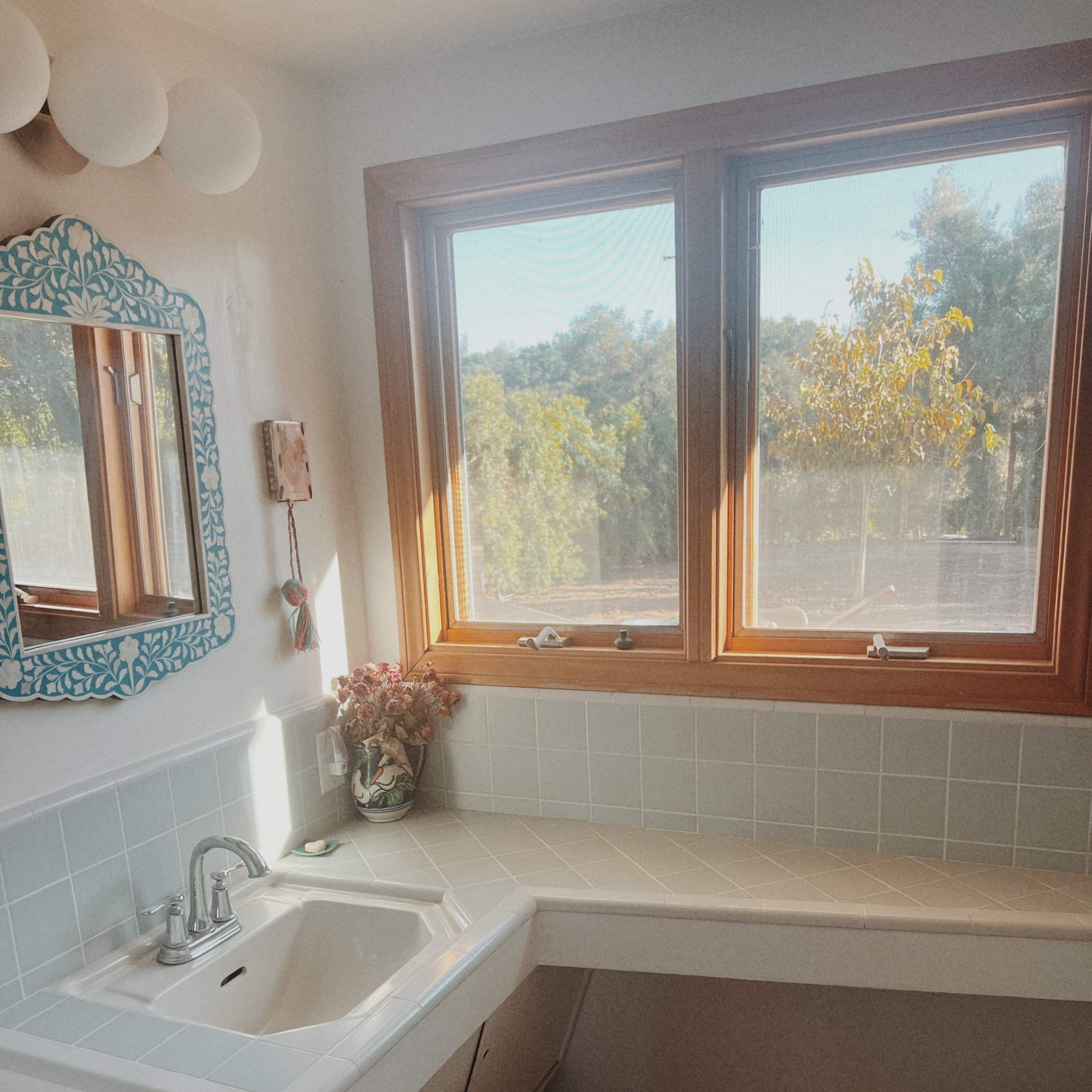 A bright bathroom featuring a sink area with a countertop, a decorative mirror, and two windows overlooking a green landscape.
