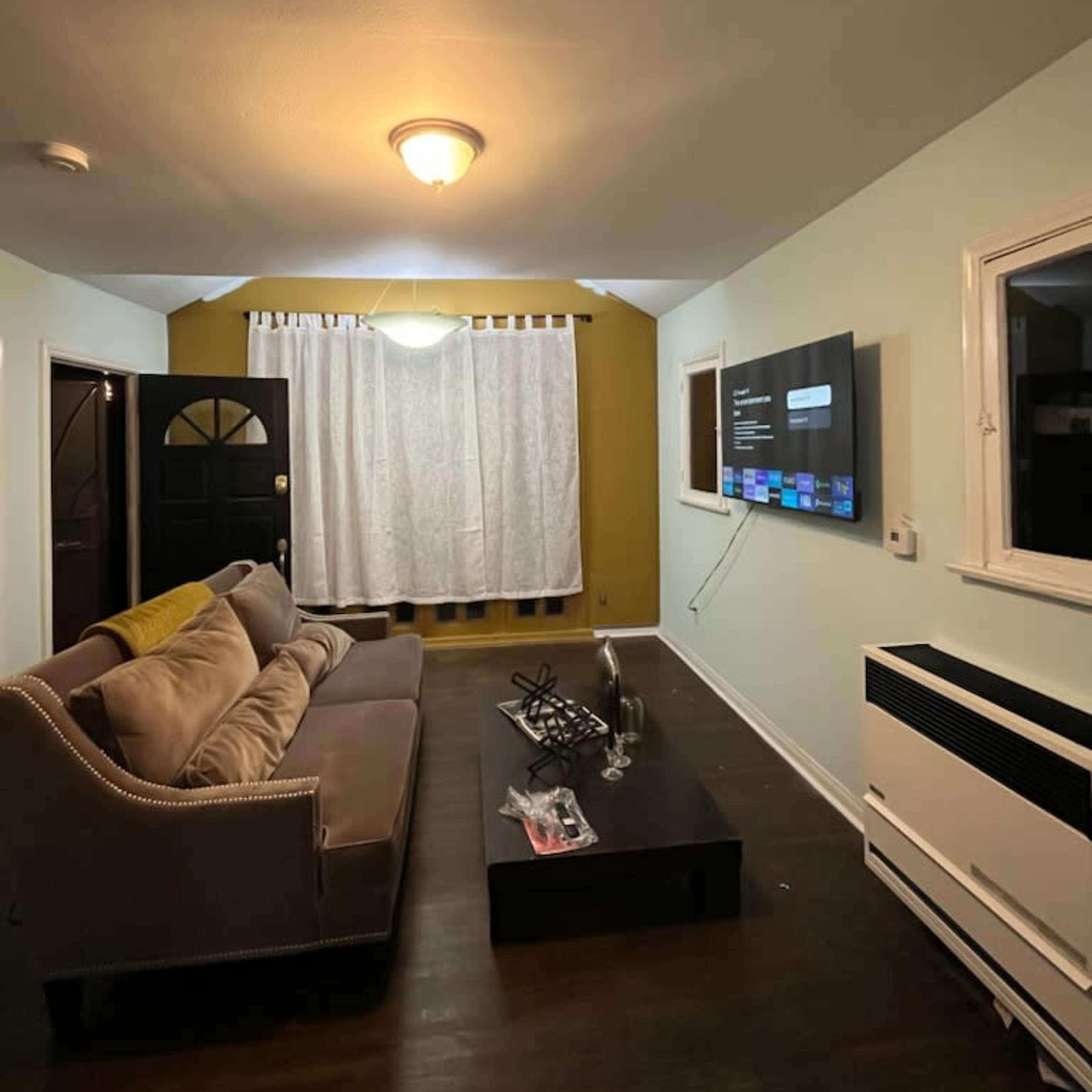 The living room features a beige sofa, a black coffee table, a mounted television on the wall, and curtains covering a window.