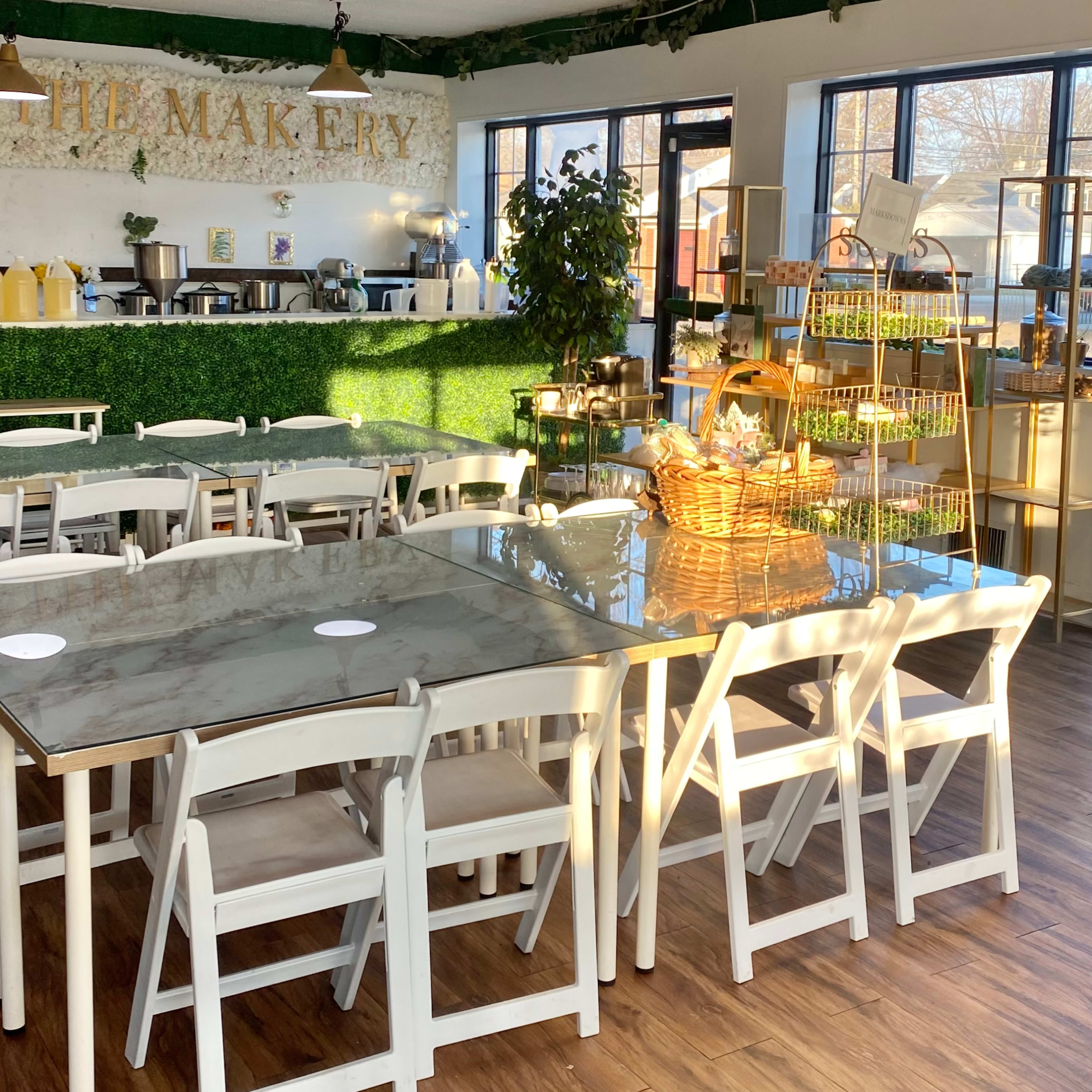 A bright café interior filled with white tables and chairs, featuring a green wall and a food display area in the background.