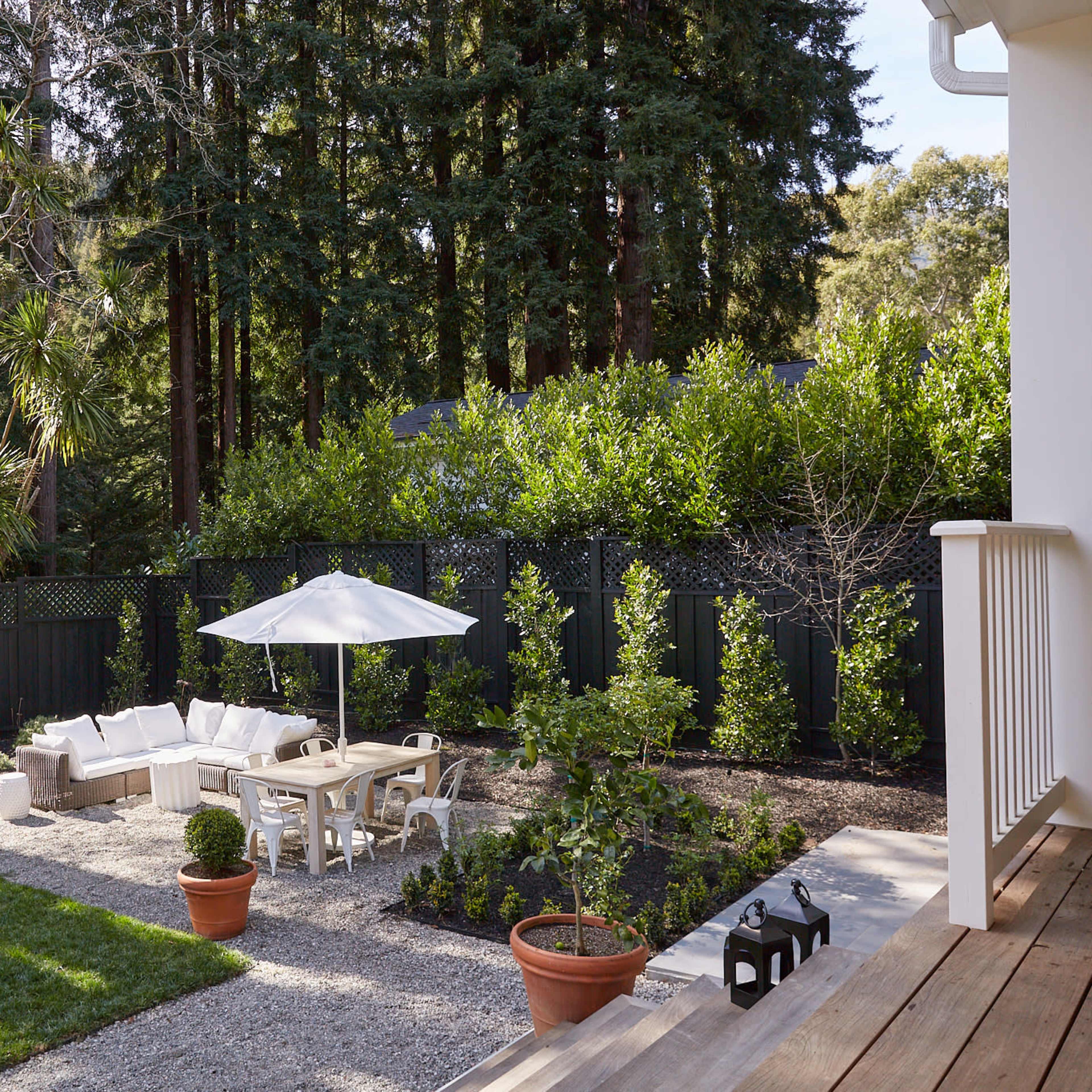 A landscaped backyard with a seating area under a white umbrella, surrounded by greenery and decorative plants.
