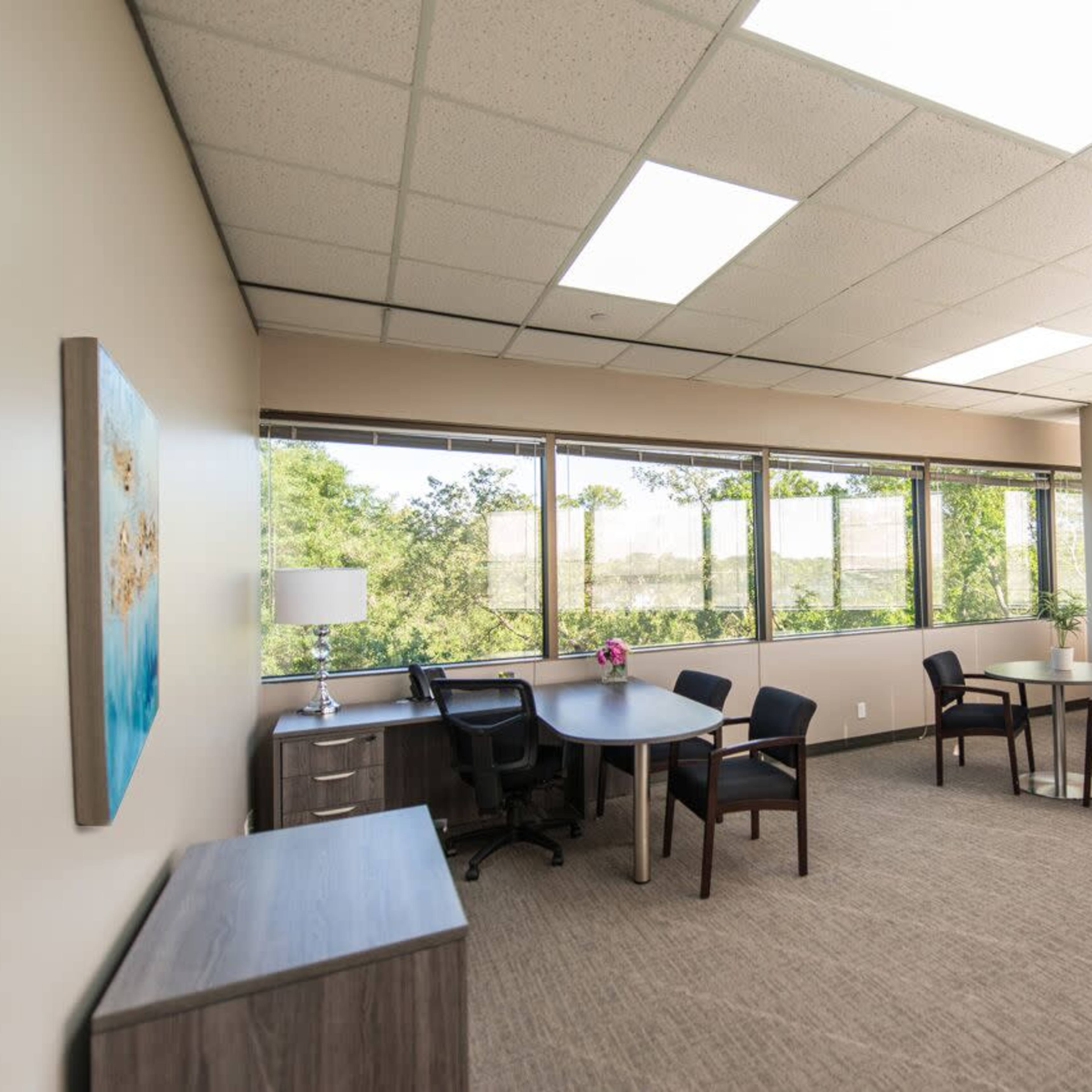 A modern office space with a round table, four chairs, and large windows allowing natural light to enter.