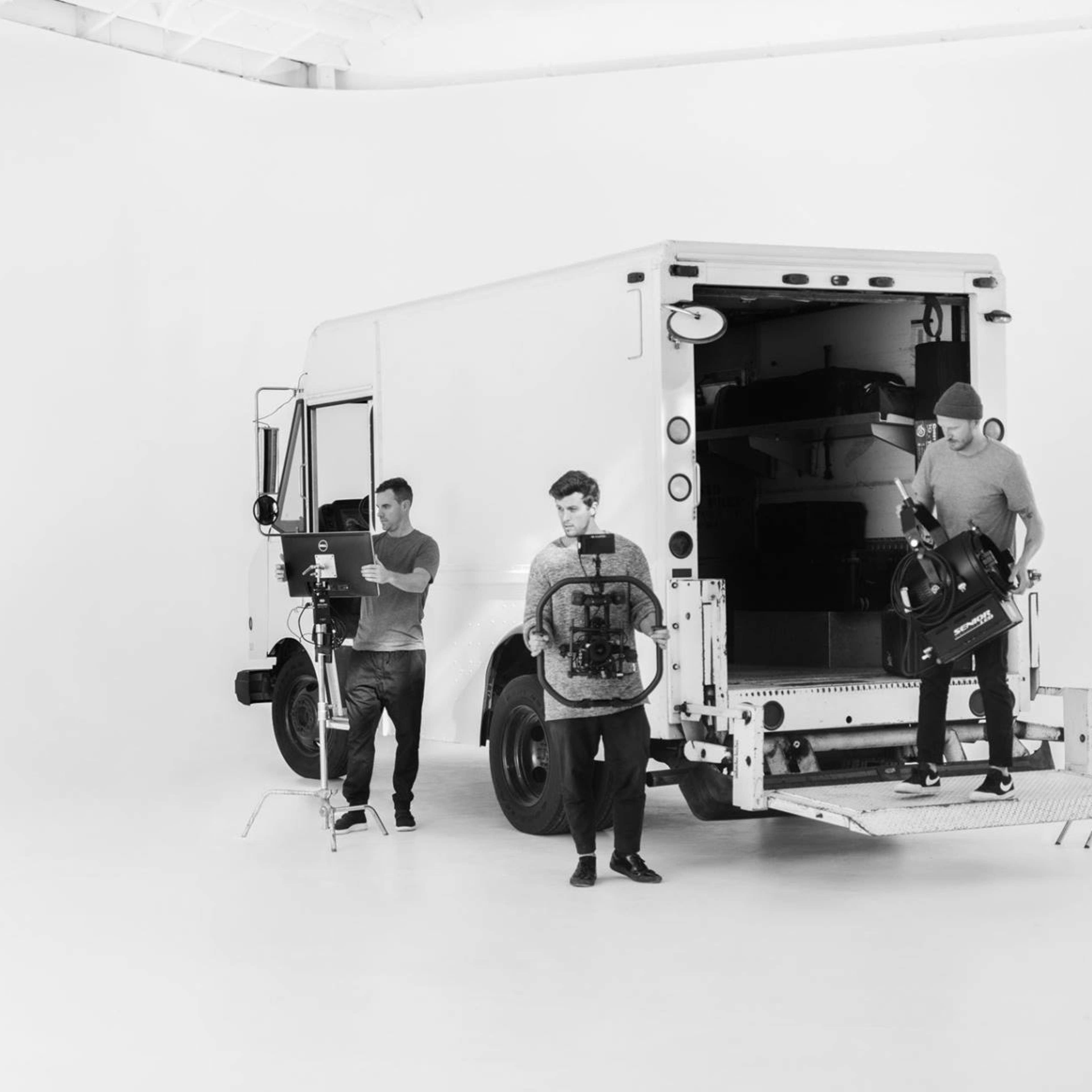 Three individuals are unloading equipment from a white delivery truck in a stark, white studio space.