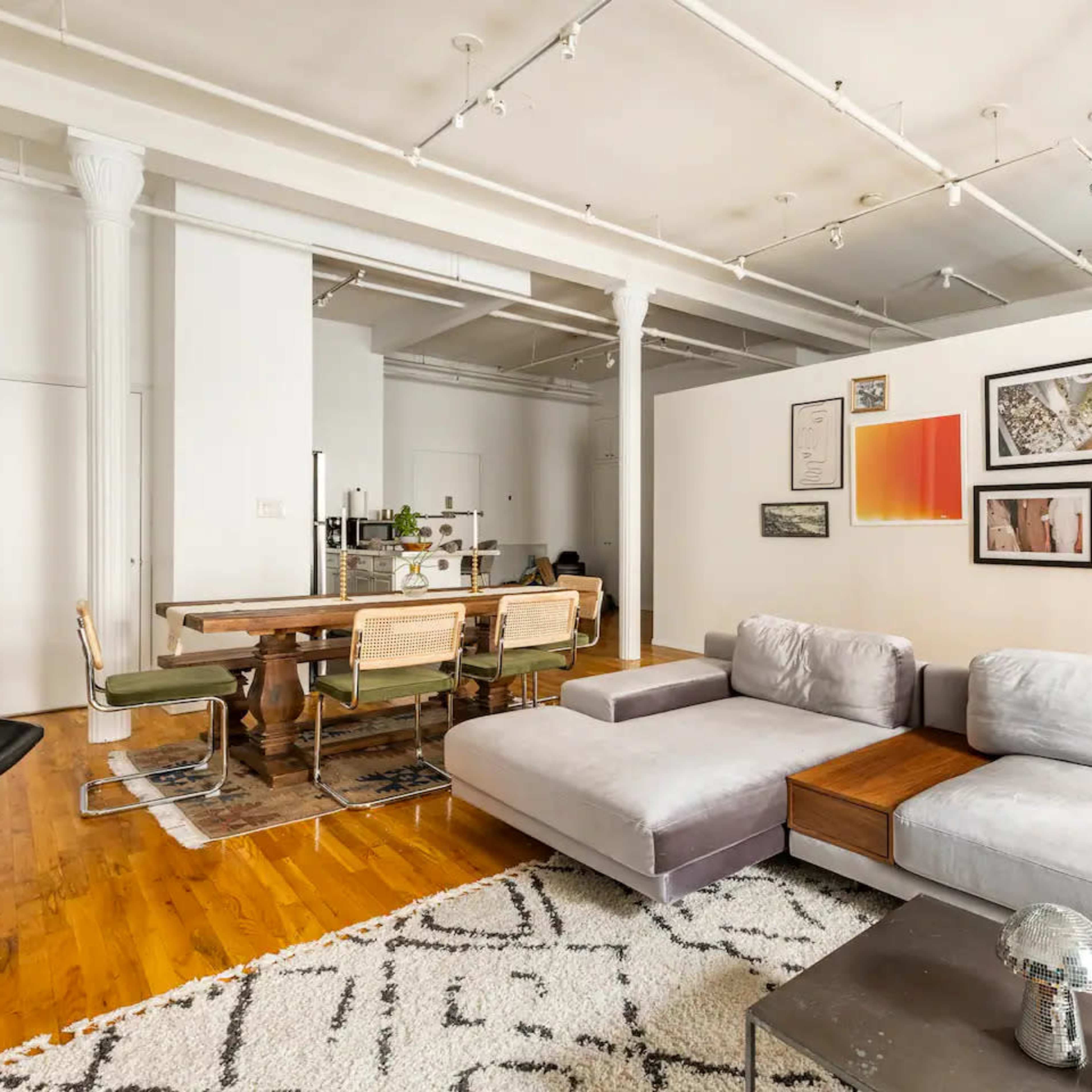 A spacious living area featuring a gray sectional sofa, a wooden dining table set for six, and various artworks displayed on the walls.