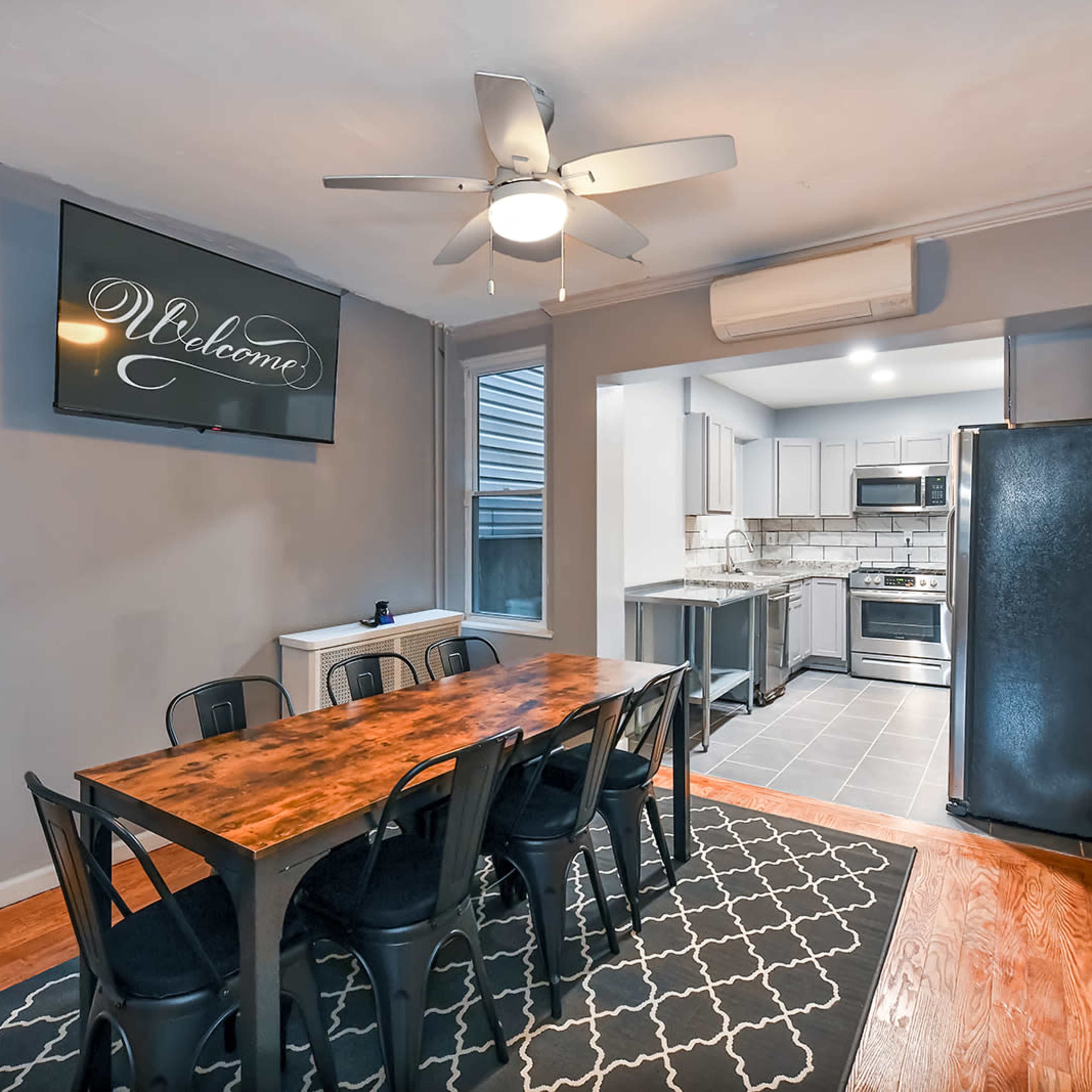 A dining area features a long wooden table with eight black chairs, a wall-mounted television, and an open kitchen space visible in the background.