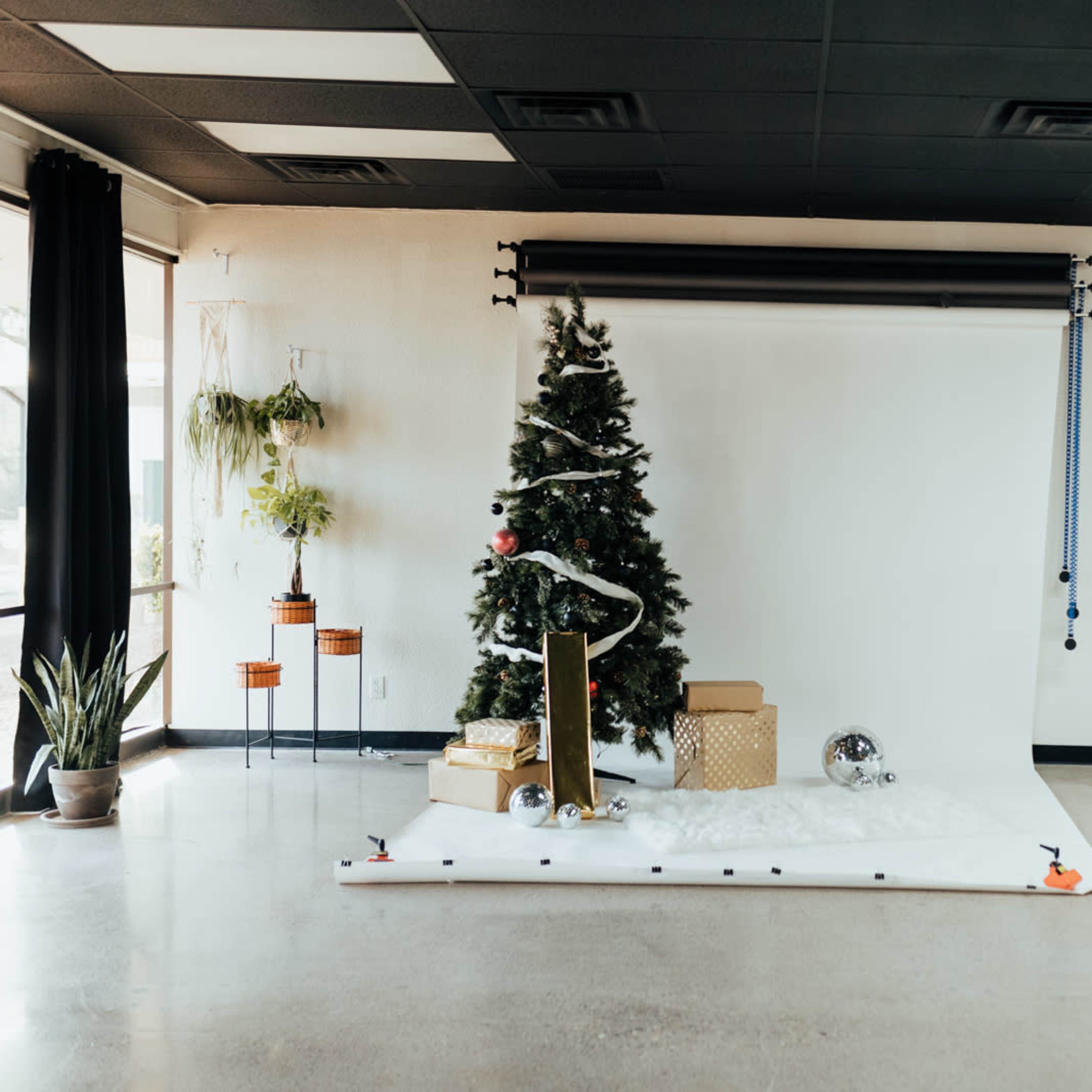 A decorated Christmas tree stands on a white backdrop with gifts and decorations arranged around it in a studio setting.