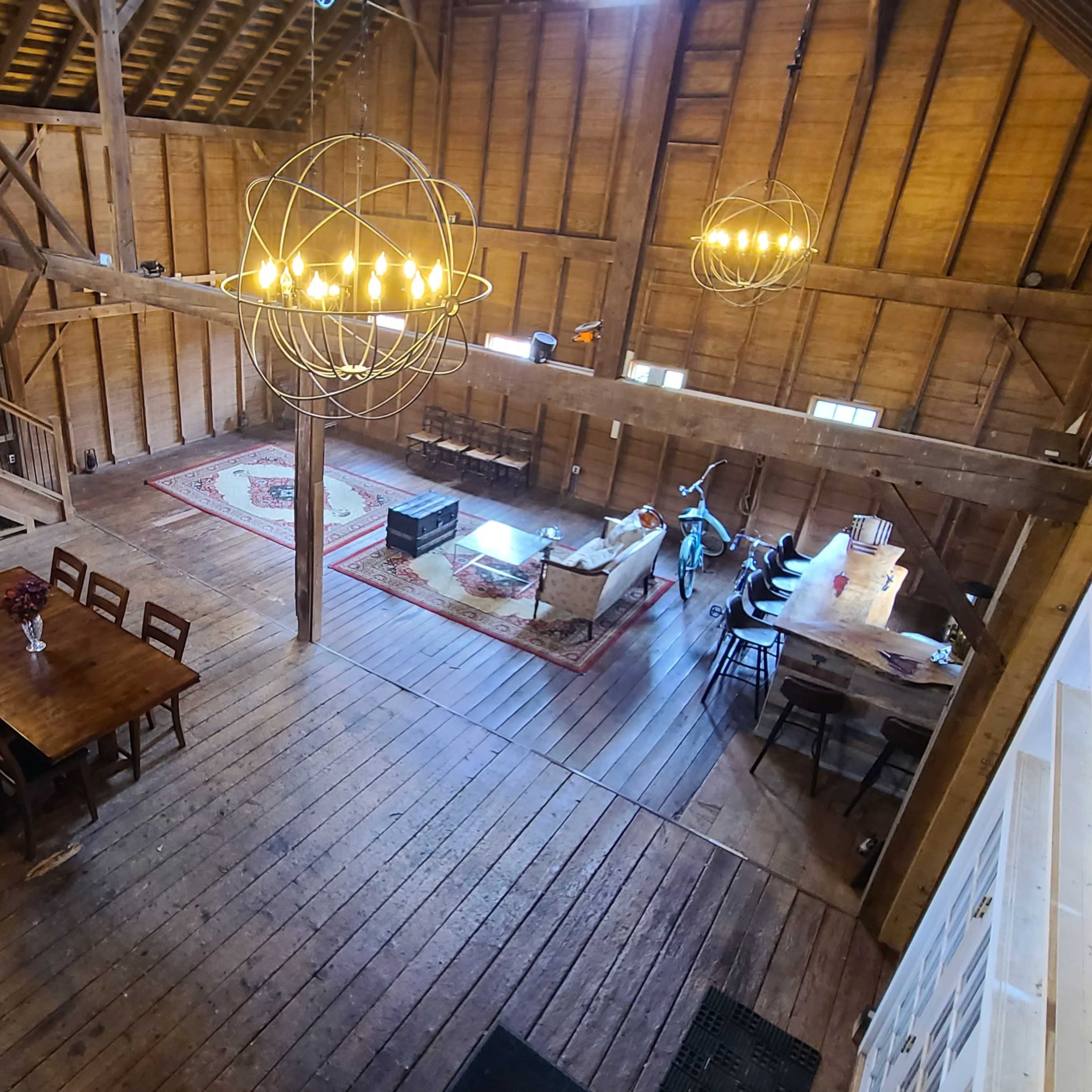 The interior of a spacious barn with wooden beams, a large dining table, and several seating areas illuminated by hanging light fixtures.