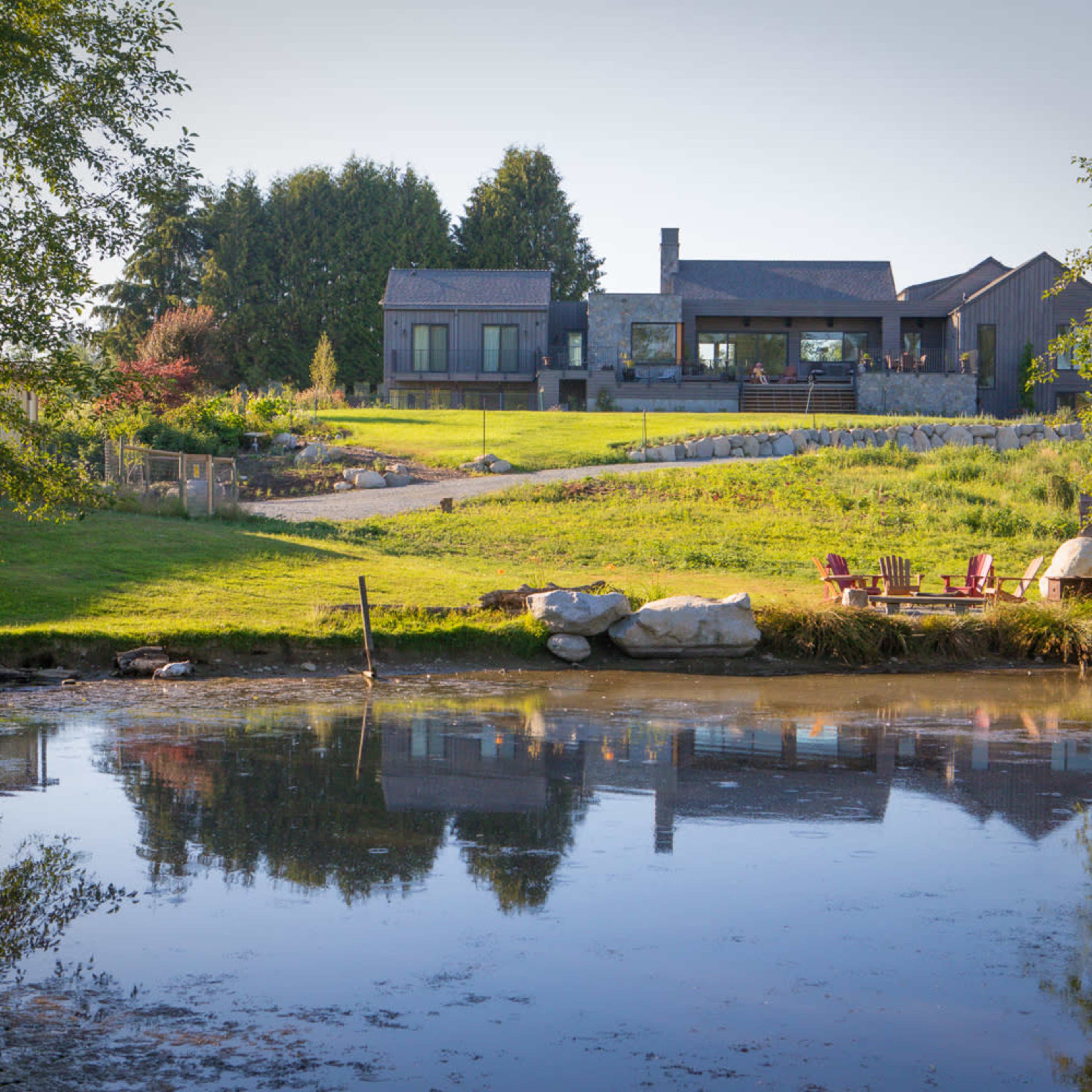 A modern house is situated near a pond, surrounded by lush greenery and trees.