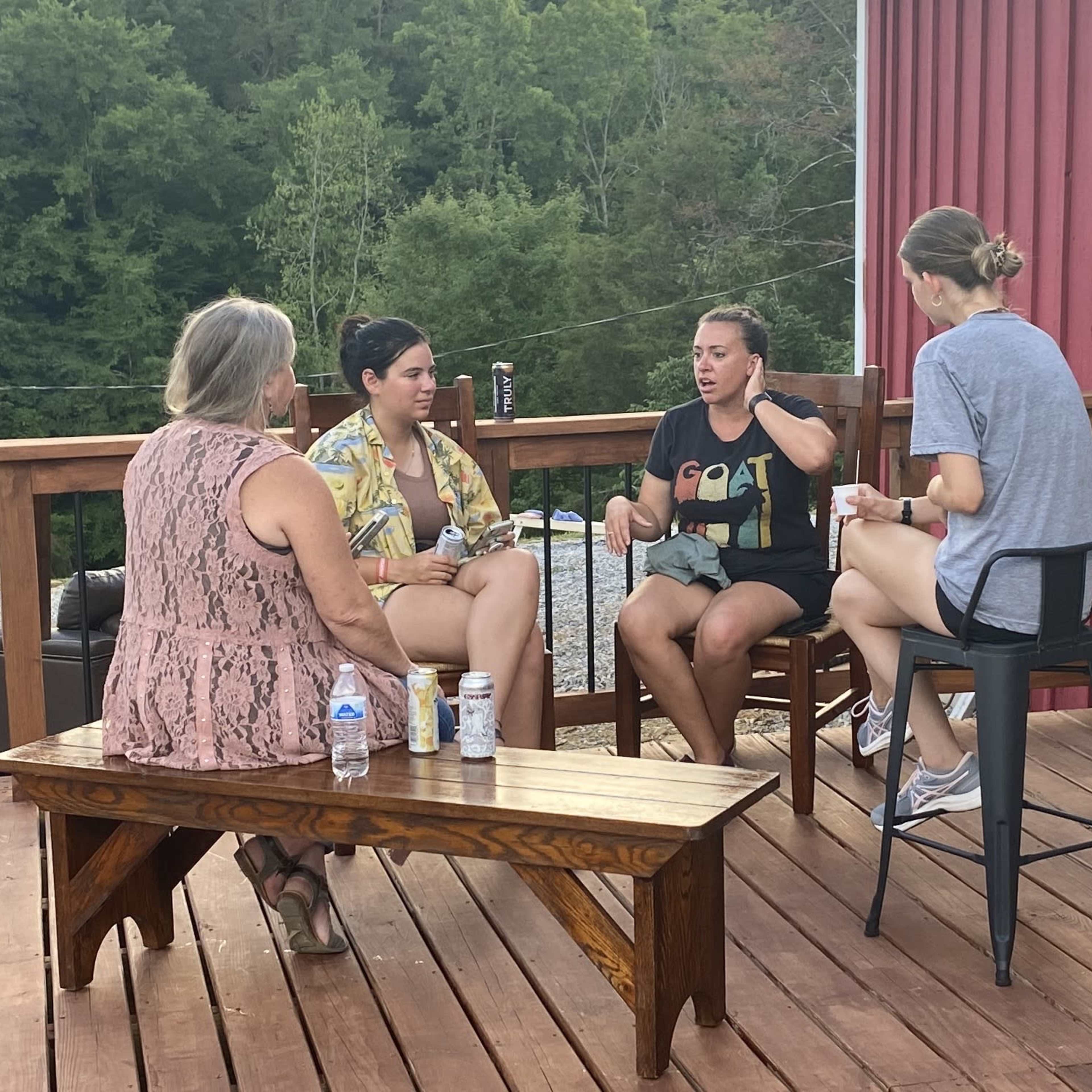 Four women sit on a wooden deck, engaged in conversation, with trees in the background.