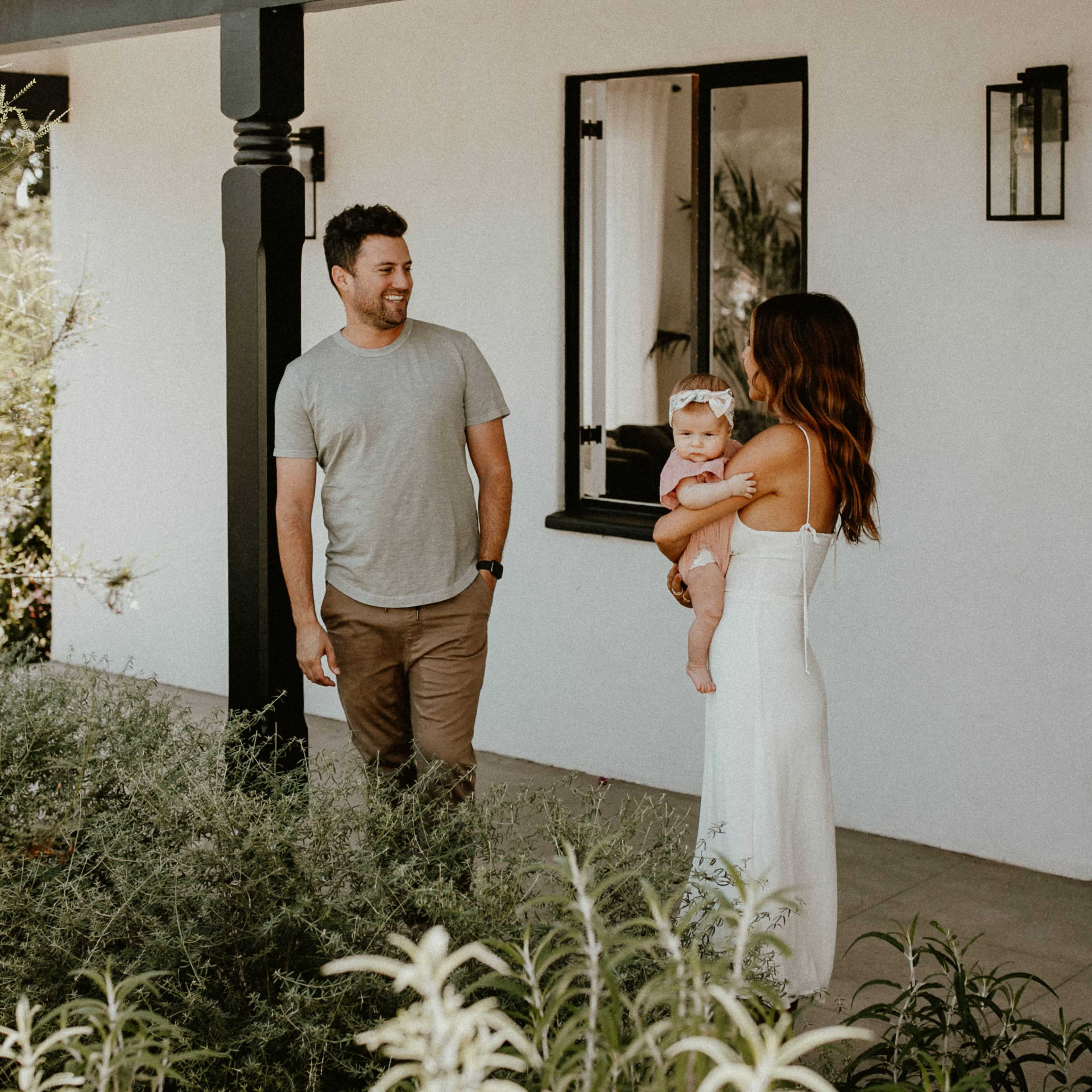 A man and a woman stand outside a house while the woman holds an infant.