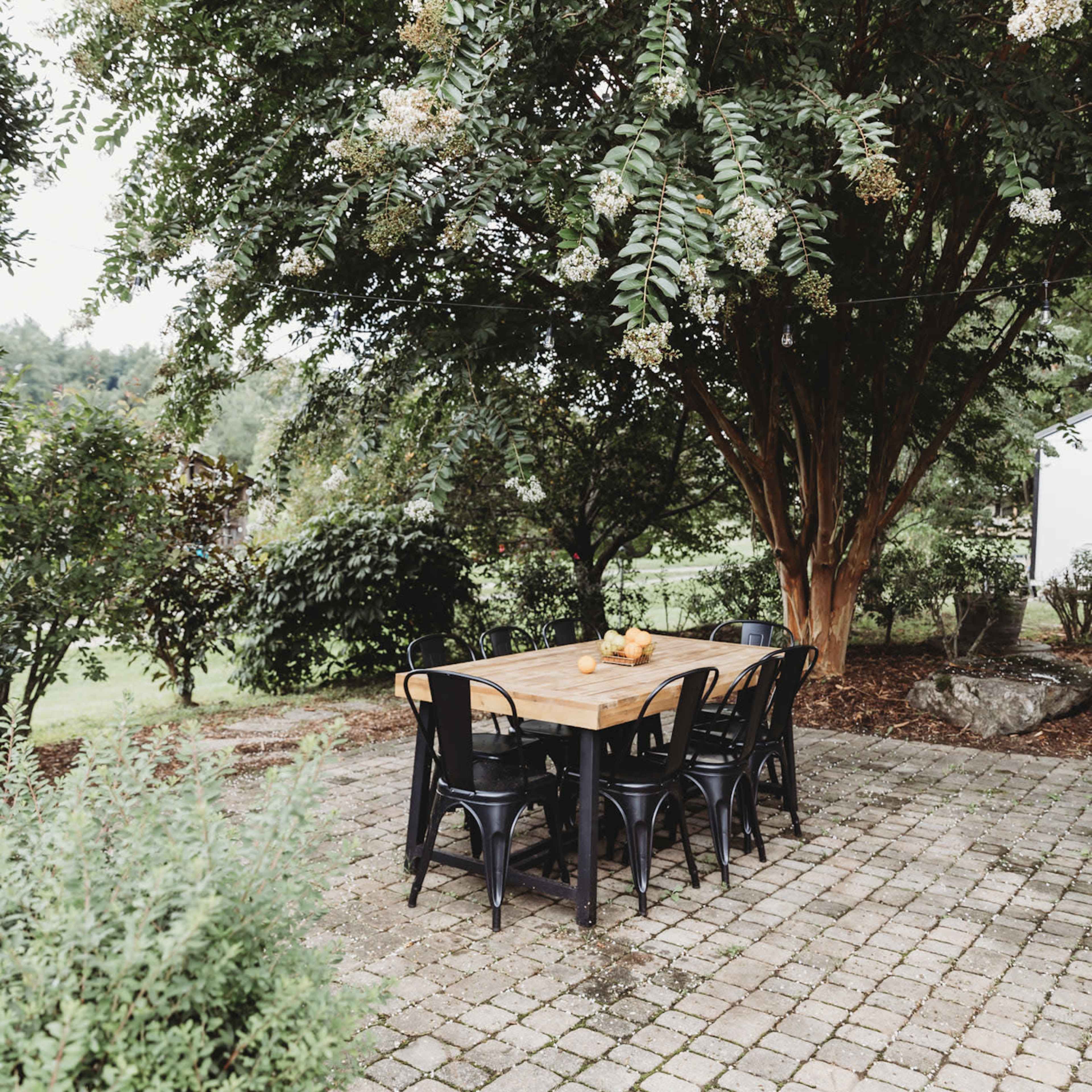 A rectangular wooden dining table with black chairs is set on a stone patio under a large tree.