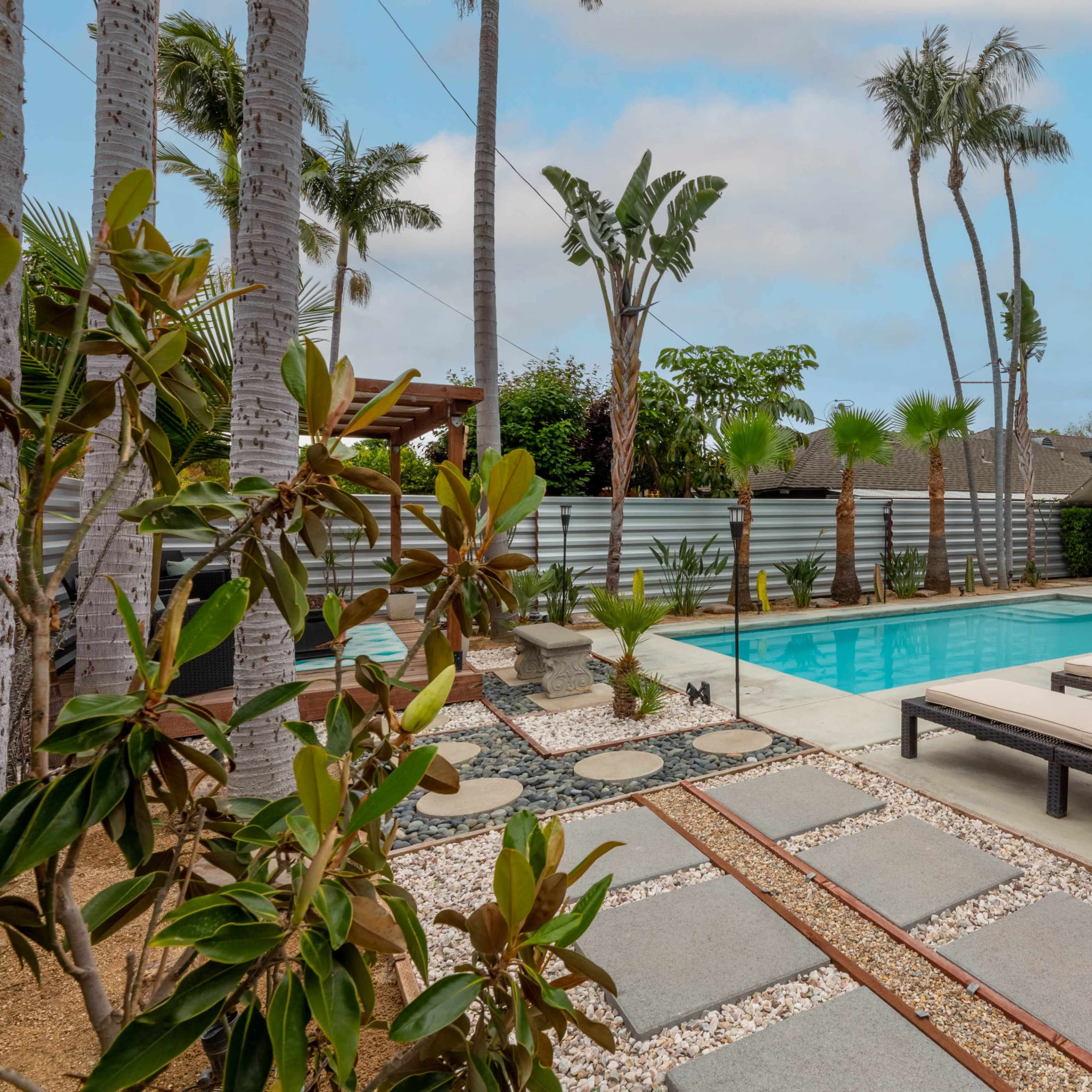 A modern outdoor space features a swimming pool surrounded by palm trees, stone pathways, and lounge chairs.