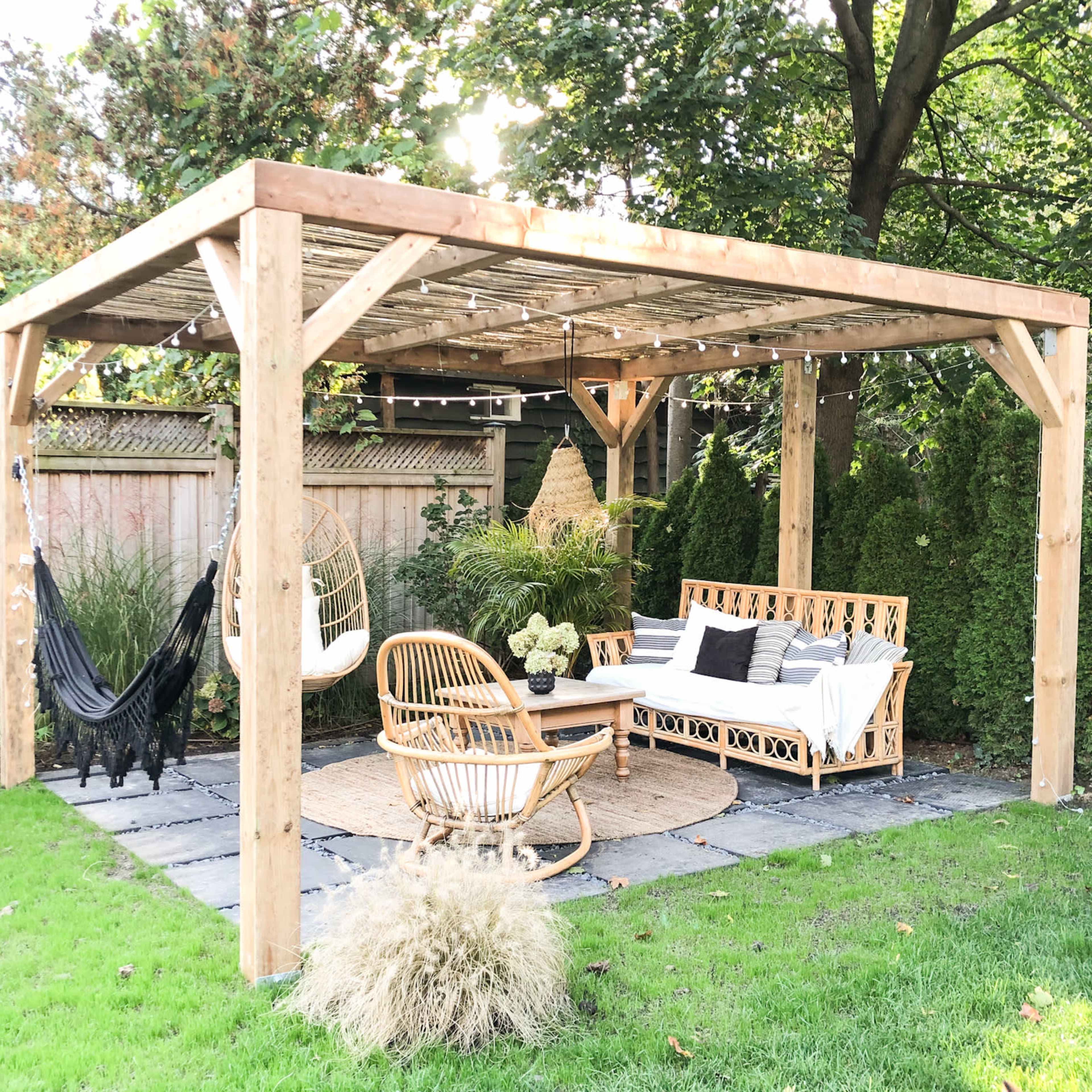 A wooden pergola over a seating area in a backyard, featuring a rattan sofa, a circular coffee table, and hanging chairs surrounded by greenery.