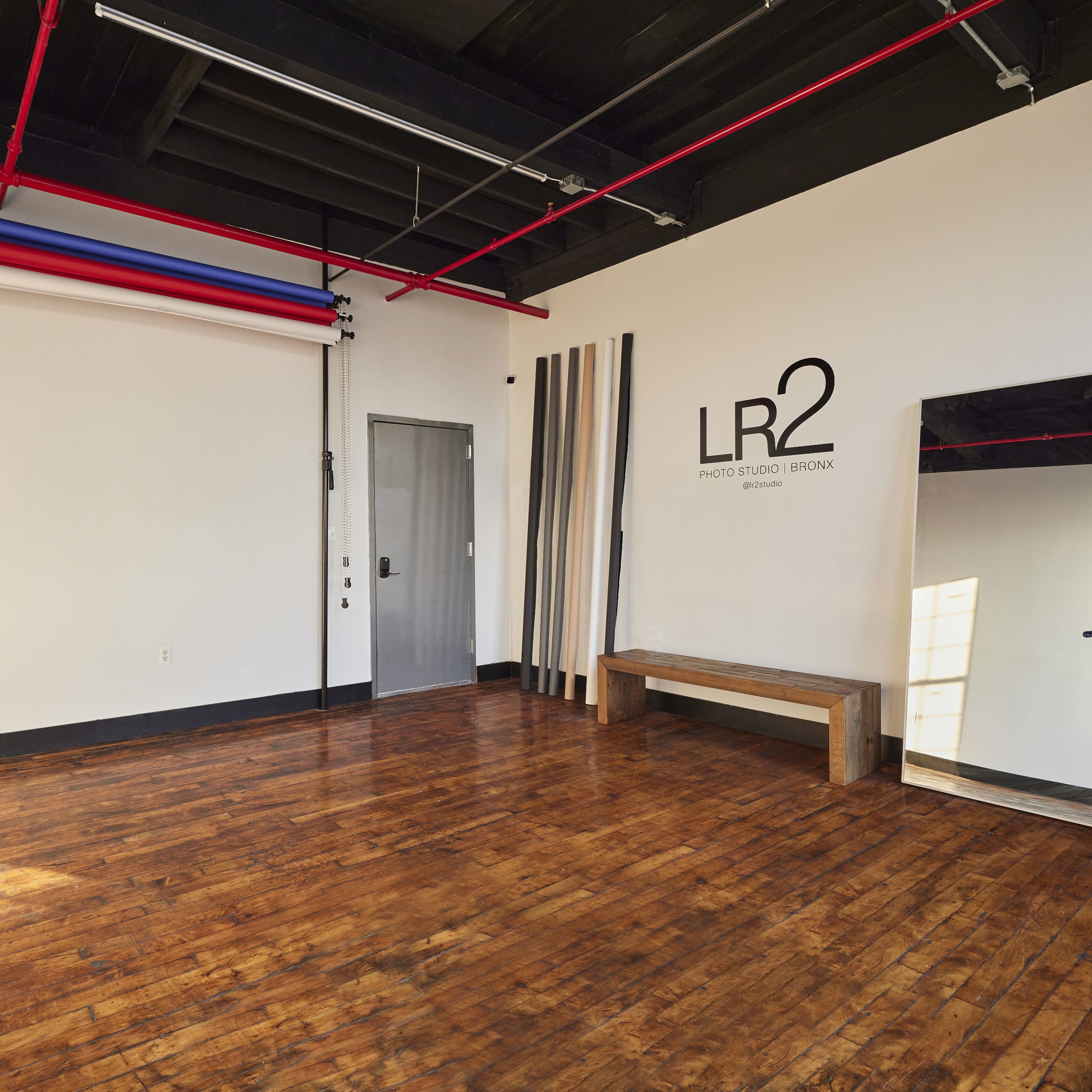 A spacious, empty studio with wooden flooring, a mirrored wall, a bench, and a few vertical props against a white backdrop.