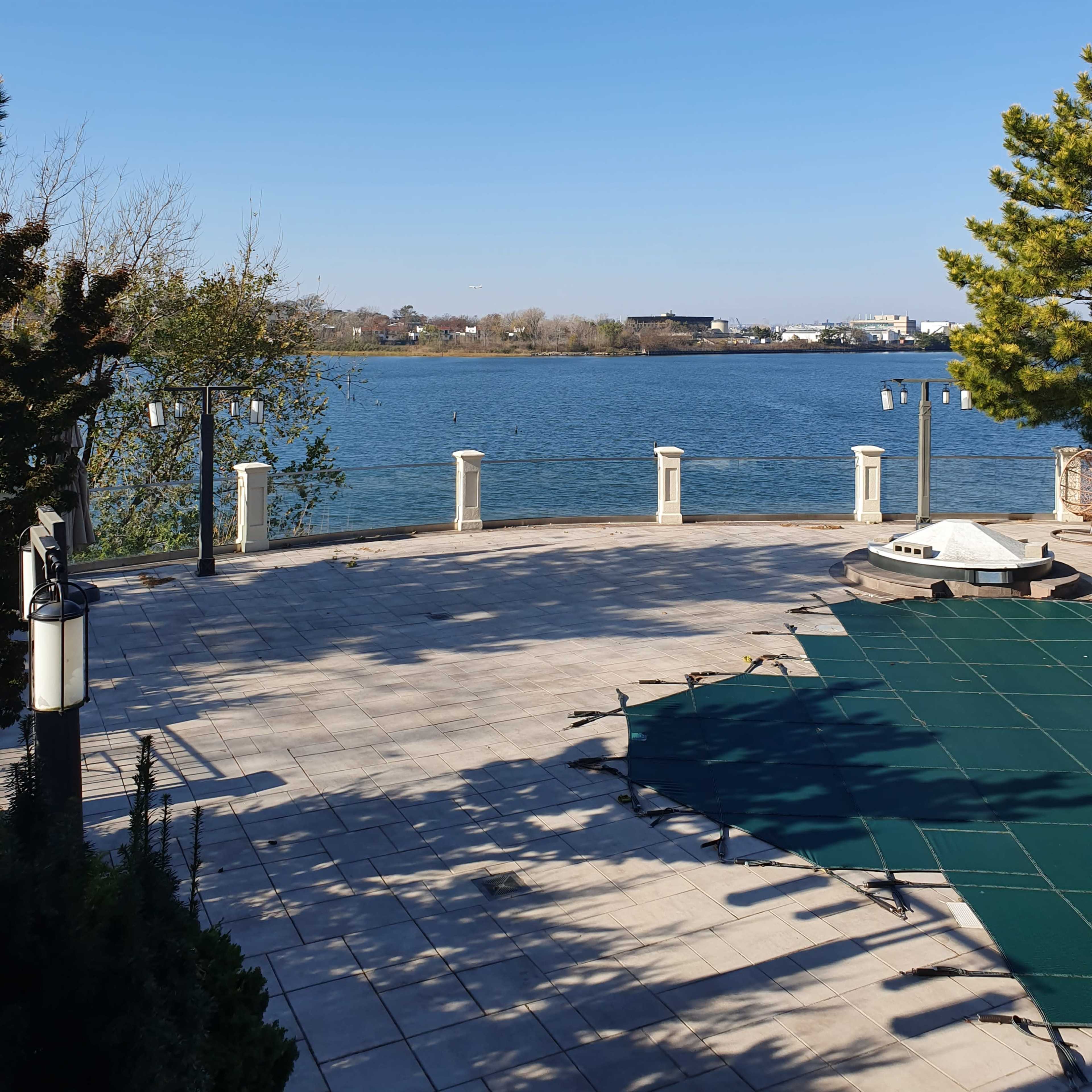 A clear view of a calm body of water bordered by a paved area and trees under a blue sky.