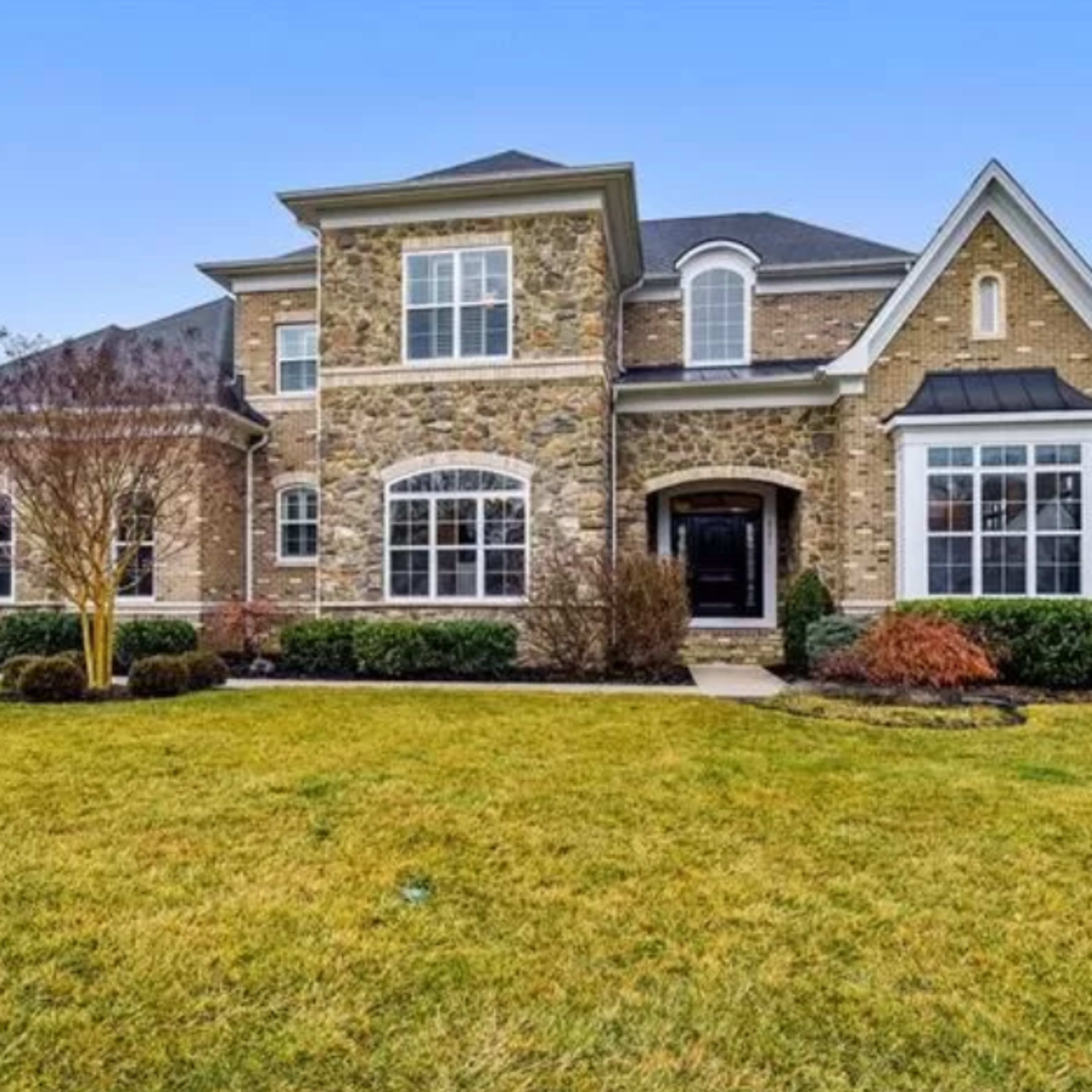 A large two-story house with stone and brick exterior is set on a landscaped lawn with neatly trimmed hedges.