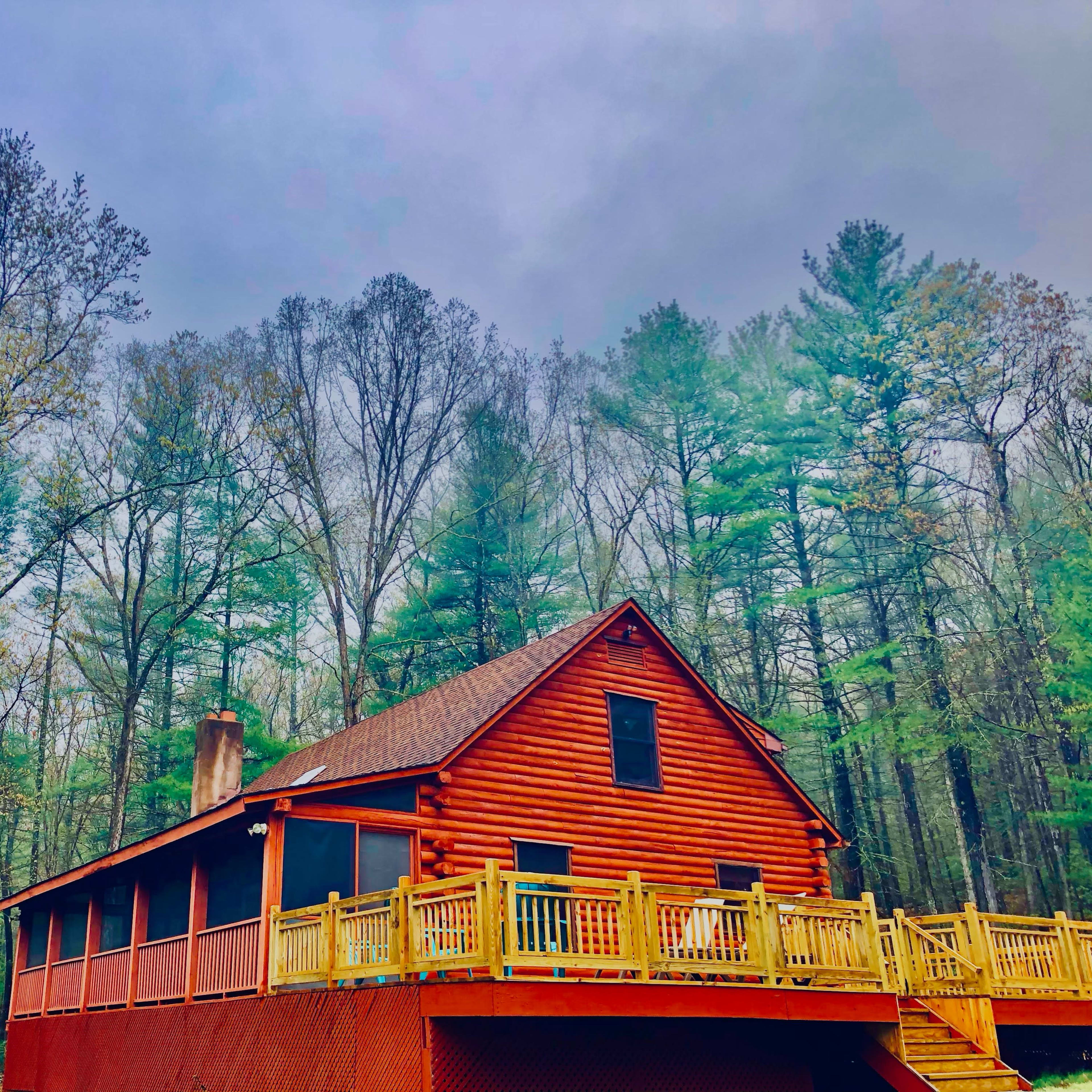 A red wooden cabin with a deck is surrounded by tall trees under a cloudy sky.