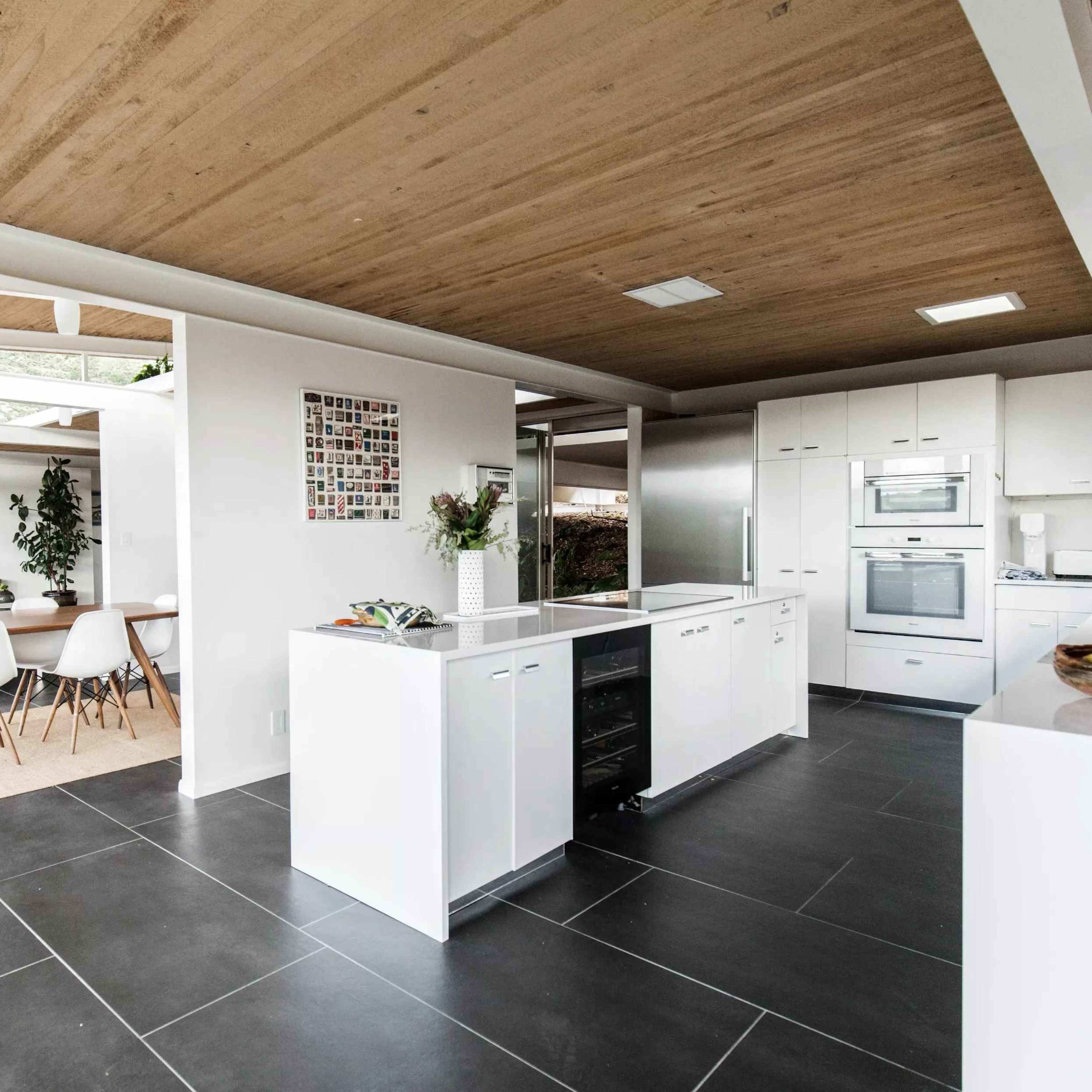 A modern open-concept kitchen and dining area with wood ceilings, sleek cabinetry, and a dining table surrounded by white chairs.