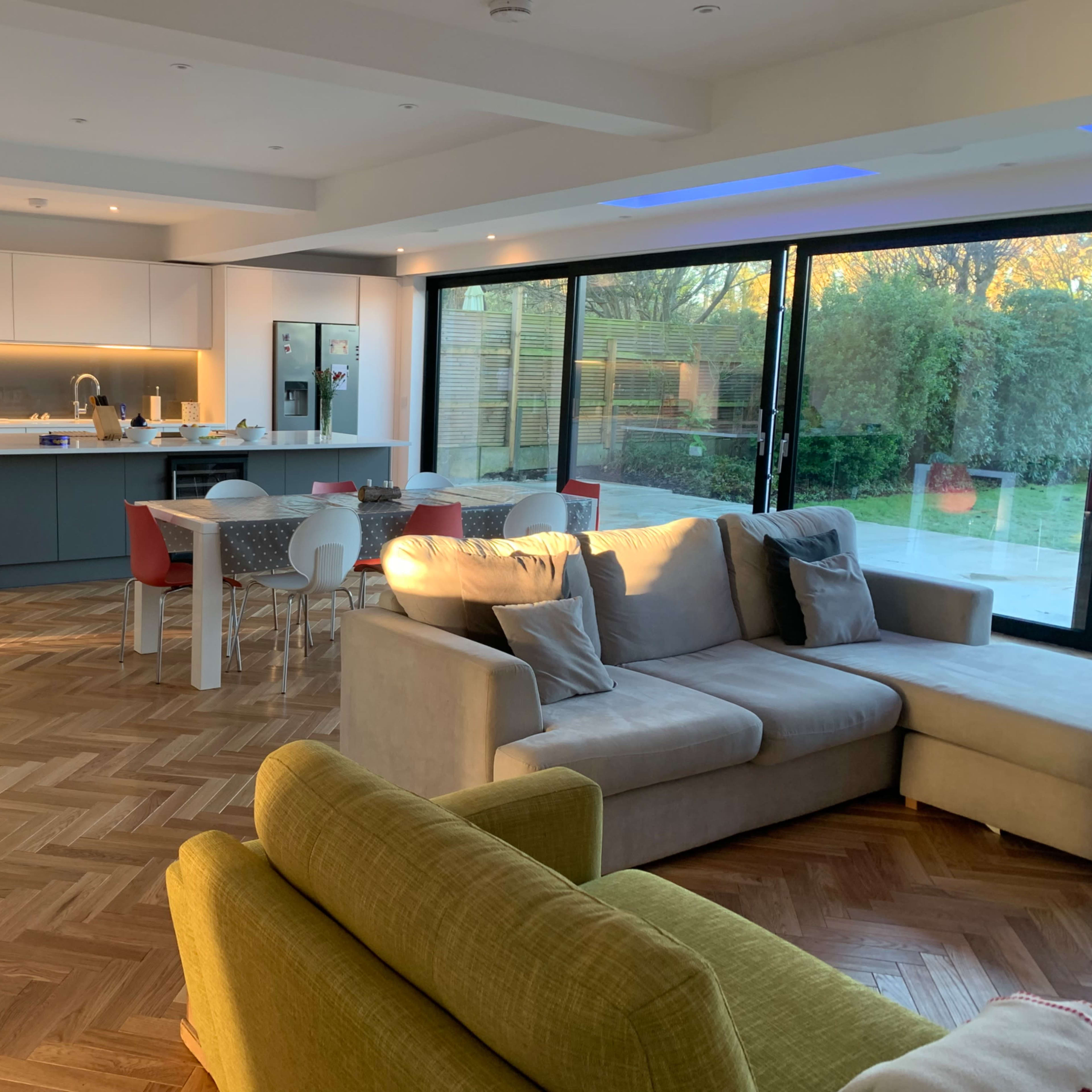 A modern kitchen and living area feature a large sectional sofa, a dining table with colorful chairs, and floor-to-ceiling windows overlooking a garden.