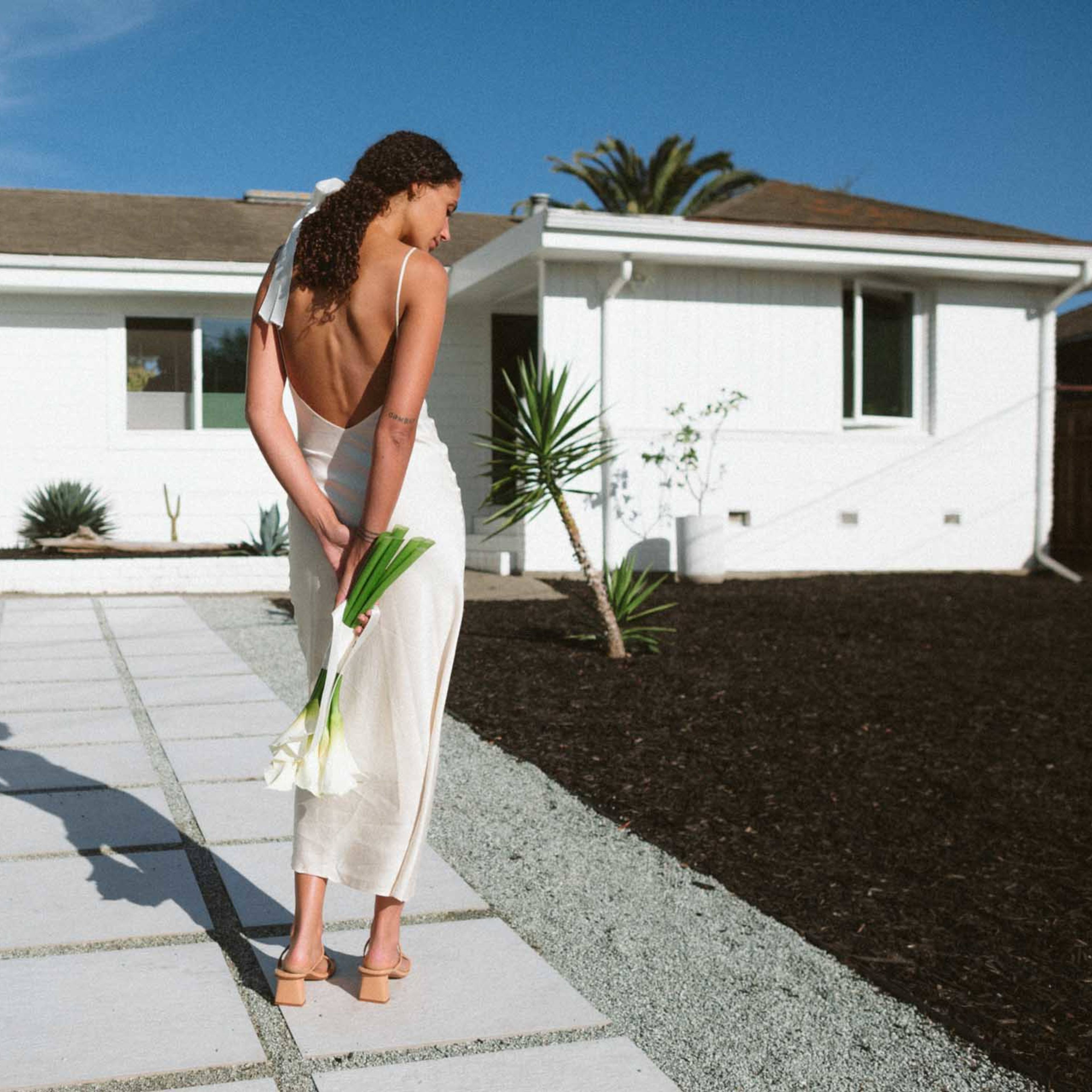 A woman stands barefoot on a stone pathway, holding white flowers behind her back in front of a white house with a blue door and palm trees.