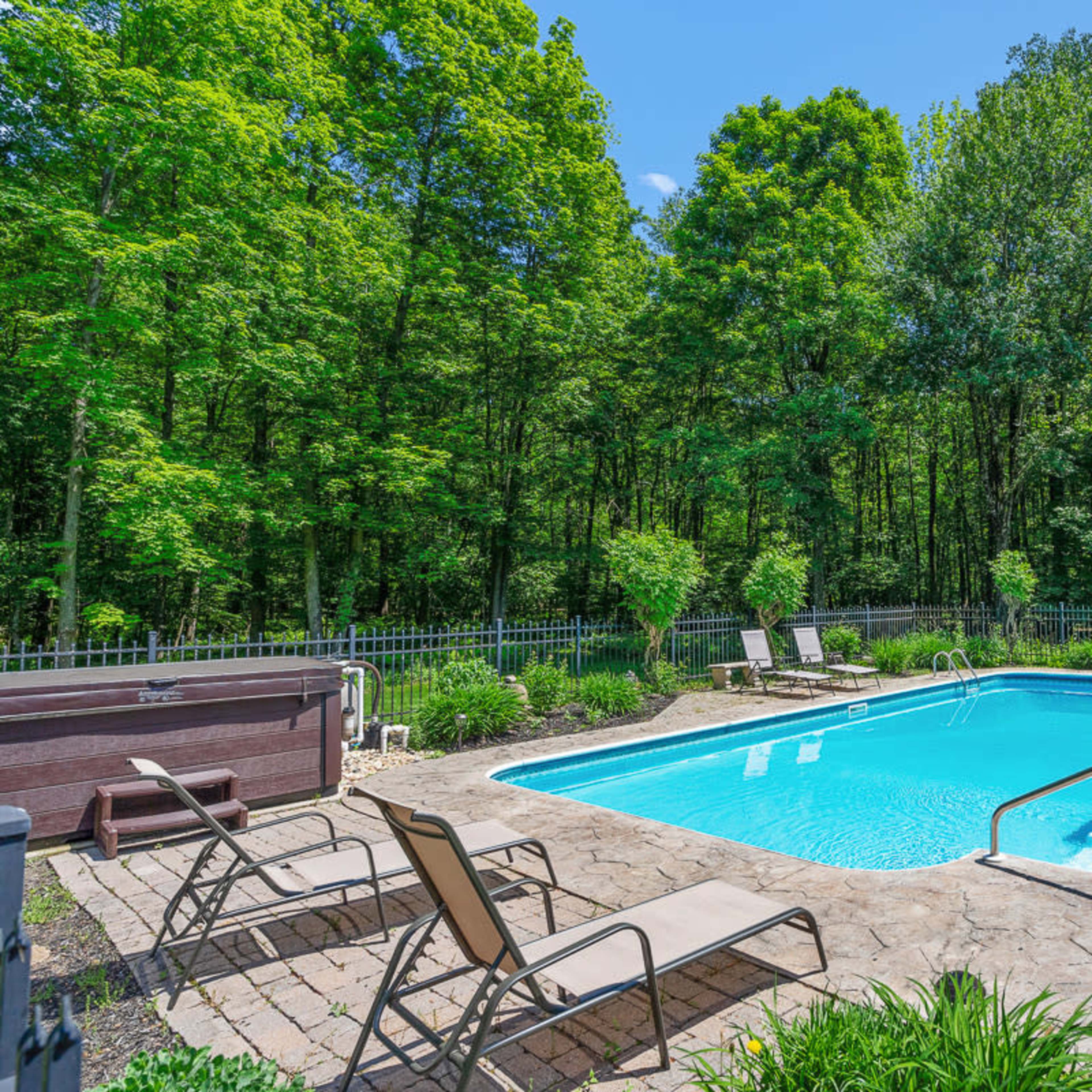 A clear swimming pool is surrounded by a stone patio and several lounge chairs, set against a backdrop of tall green trees.