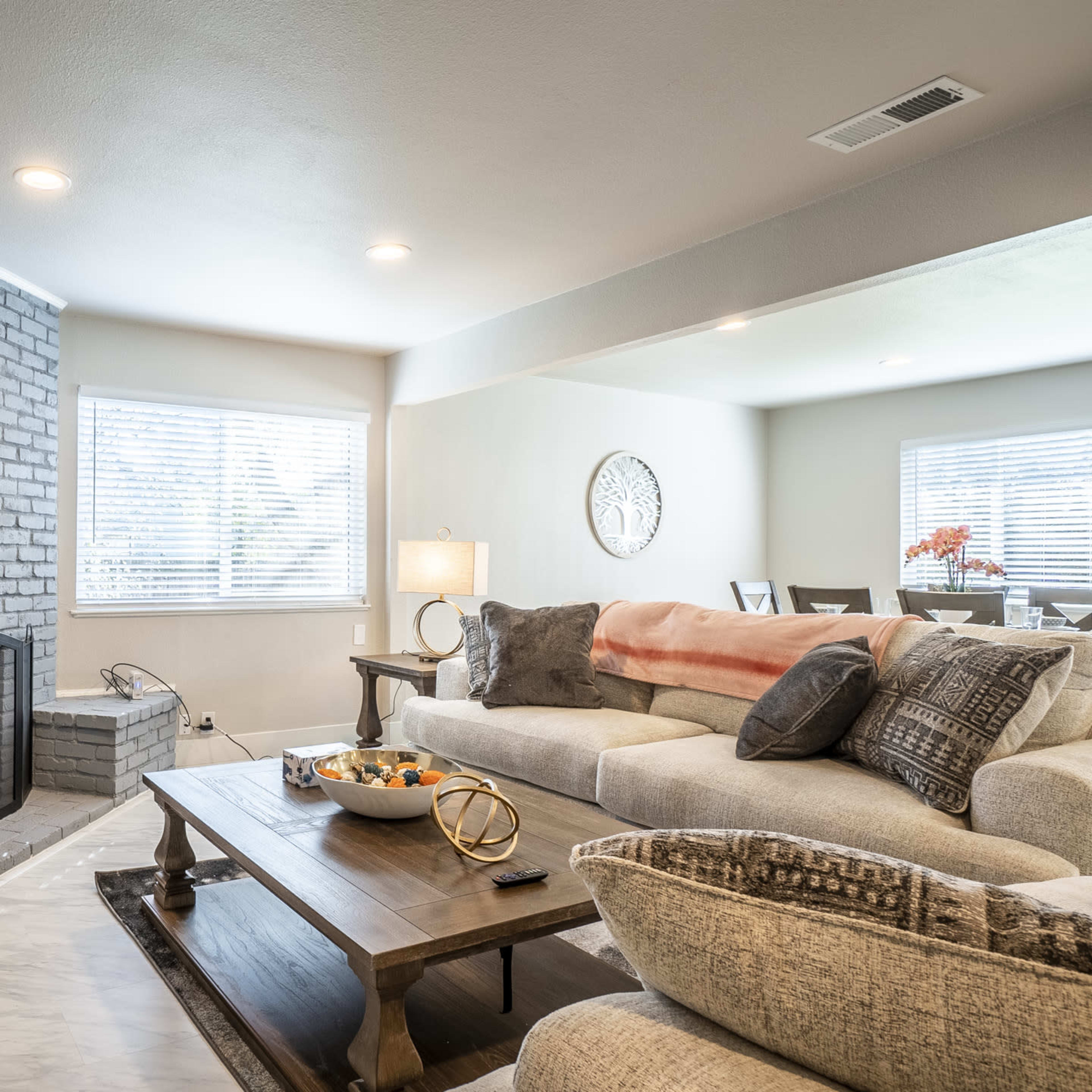 A living room featuring a gray-brick fireplace, a beige sectional sofa, a wooden coffee table with decorative items, and a dining area in the background.