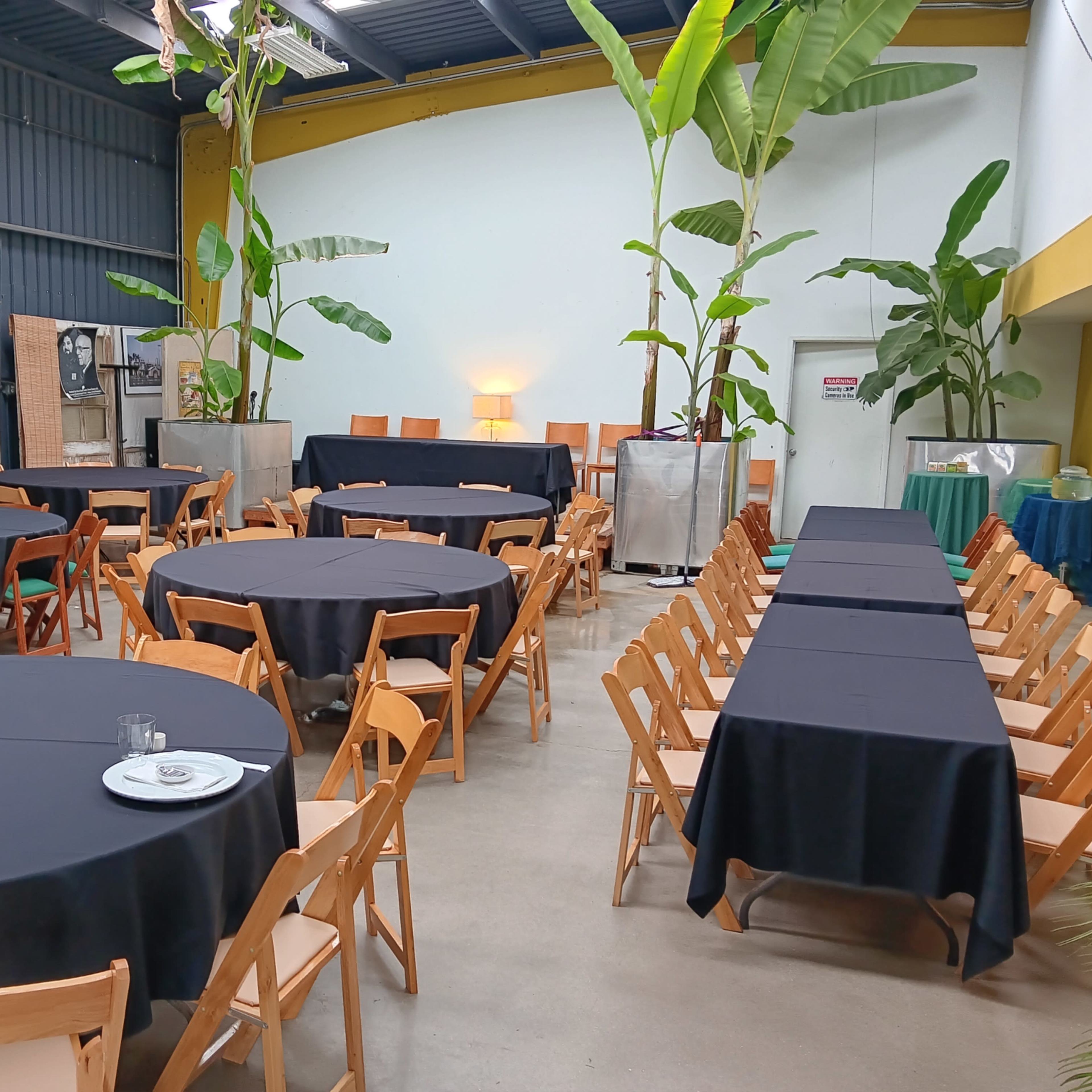 A spacious indoor venue features several round and rectangular tables covered with black tablecloths, surrounded by wooden chairs and decorative plants.