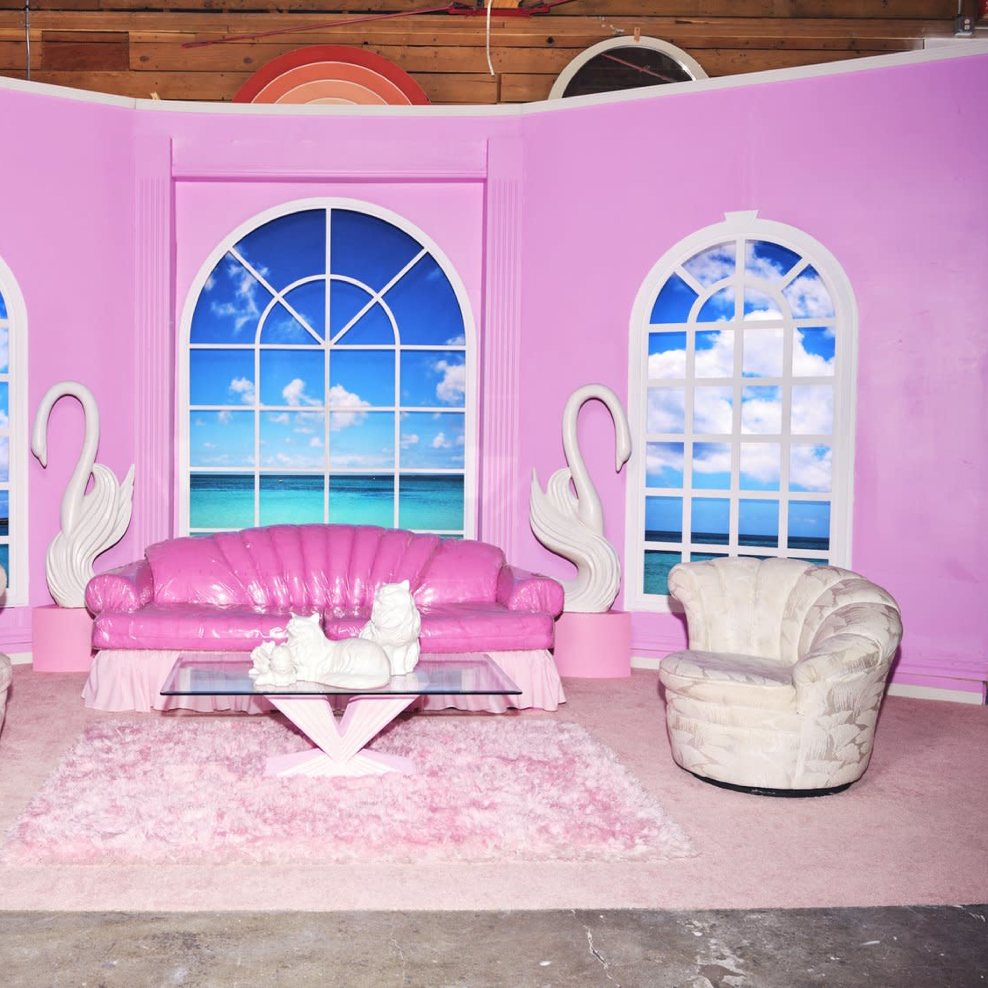 The scene features a pink room with large arched windows showing a beach view, furnished with a pink sofa, two white chairs, and a glass coffee table on a fluffy area rug.