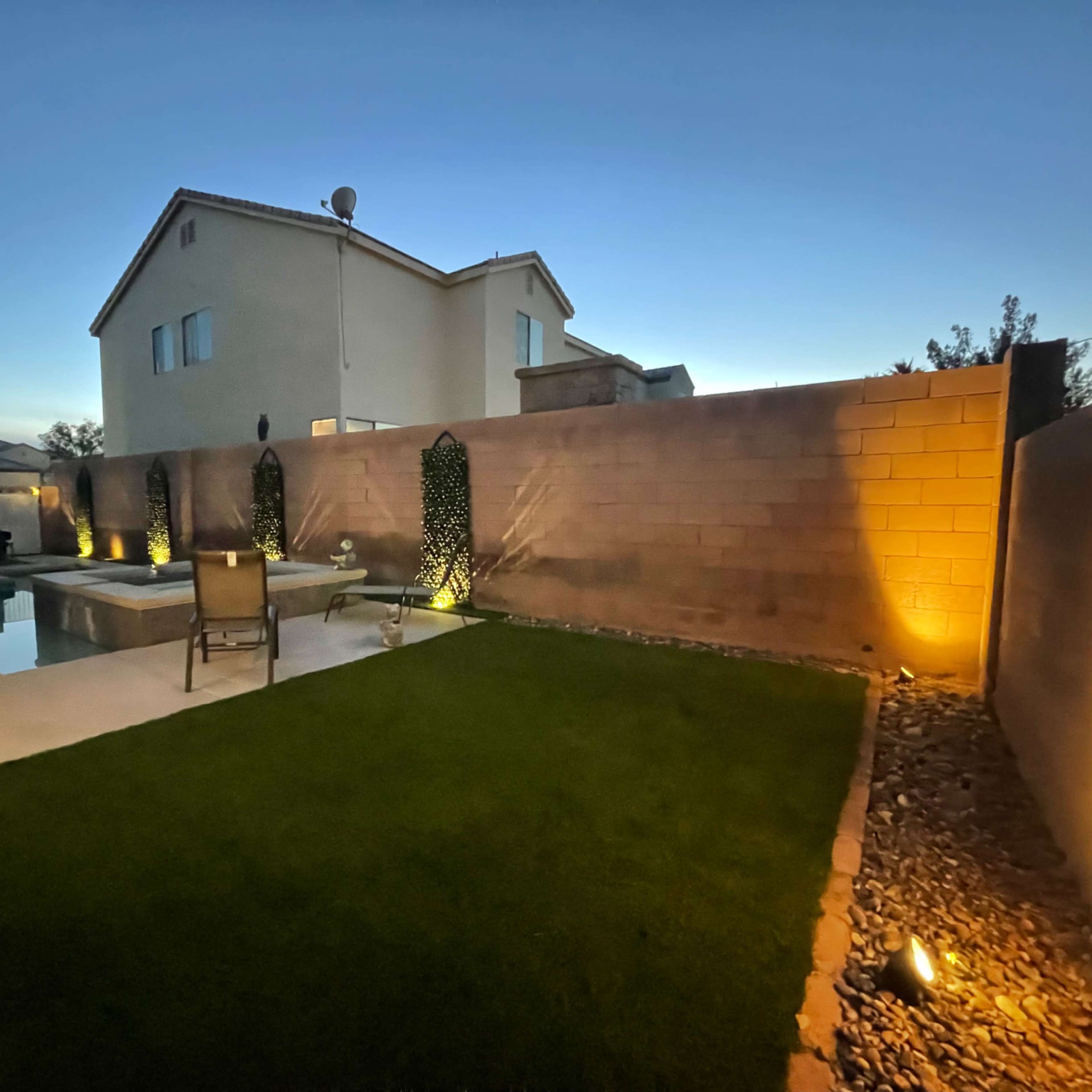 A backyard at twilight, featuring a lawn, a pool, and illuminated plants against a wall.