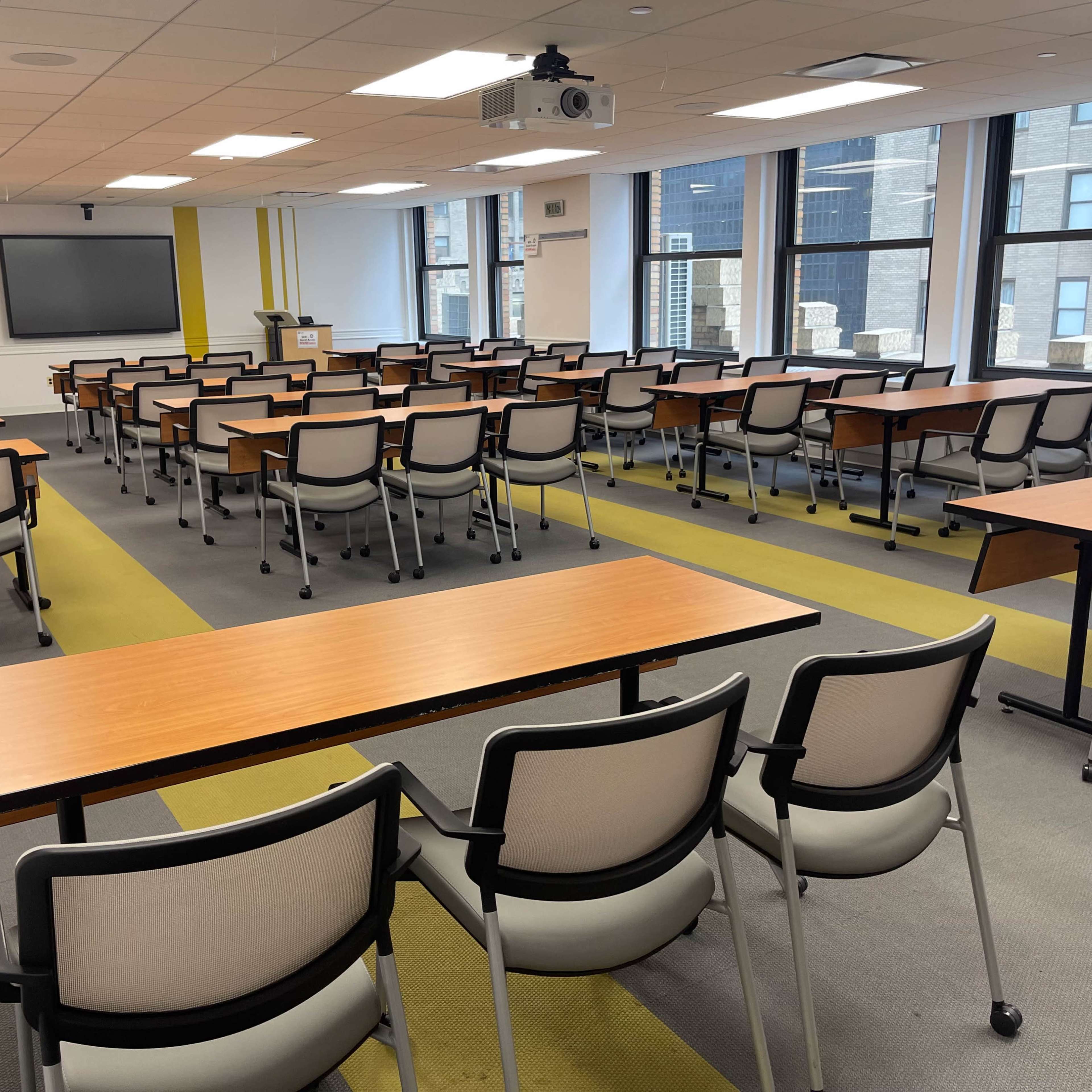 A spacious classroom is set up with rows of tables and chairs, facing a projector and screen at the front.