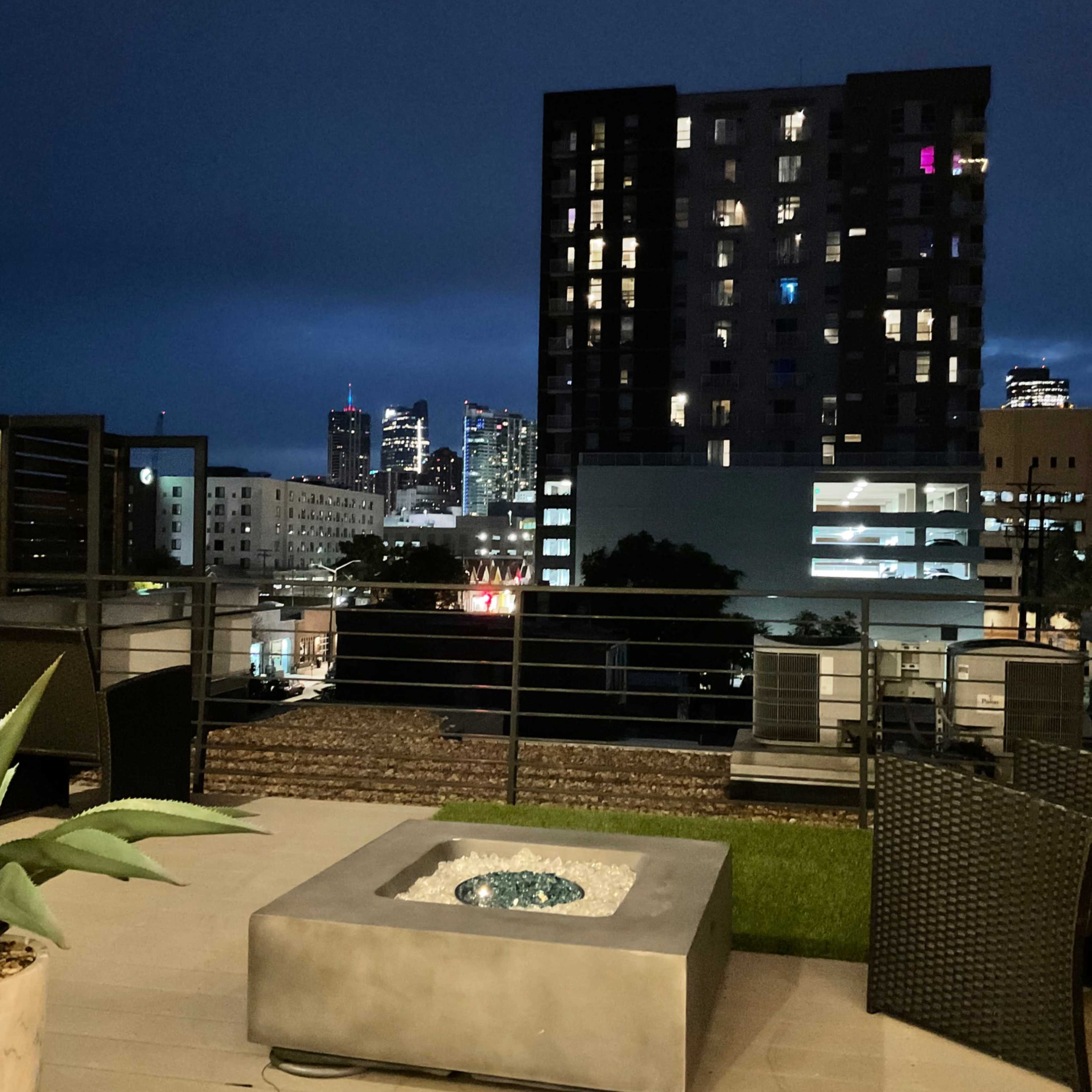 A rooftop patio at night with a modern fire pit, overlooking a city skyline highlighted by illuminated buildings.