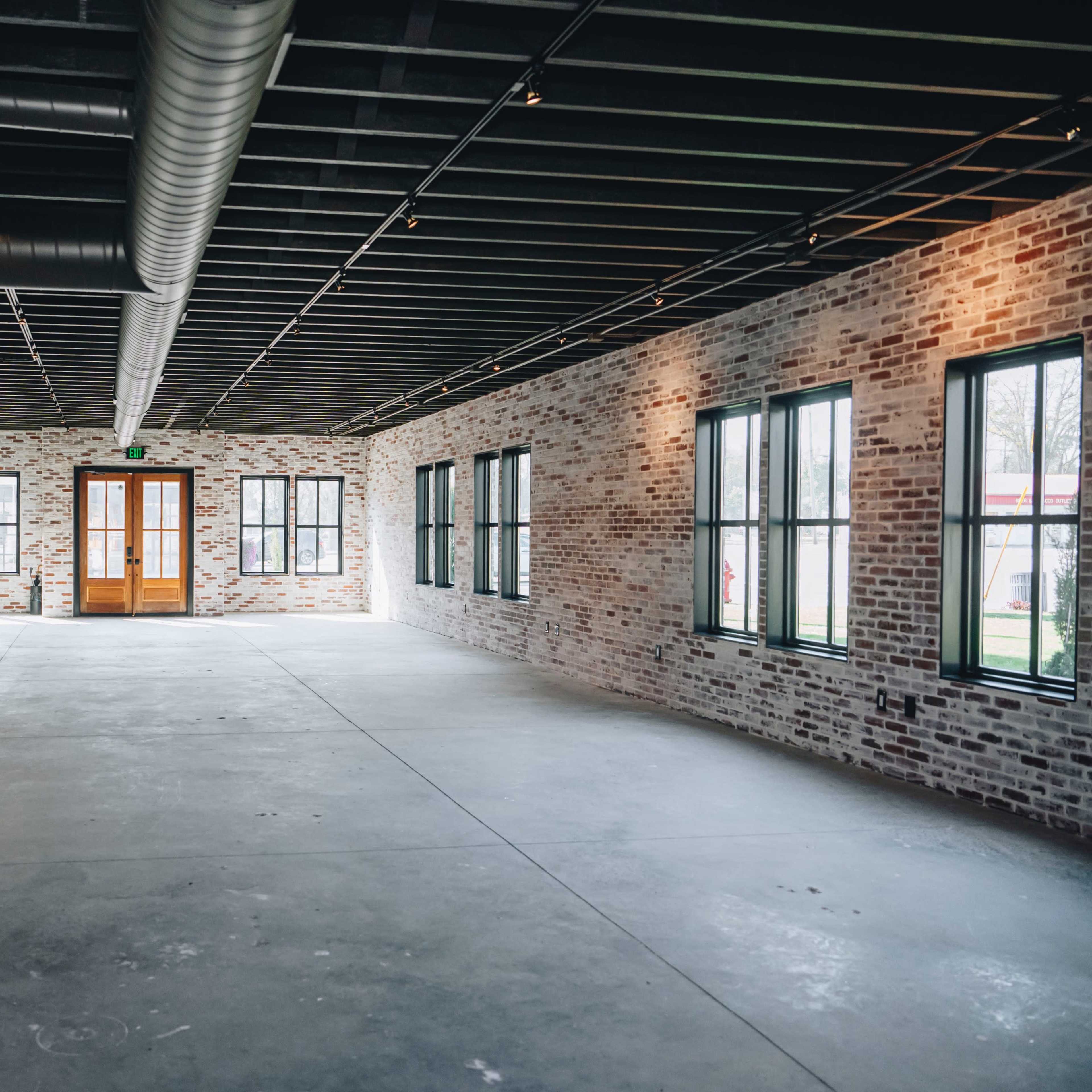 An empty interior space with exposed brick walls, large windows, and a concrete floor.